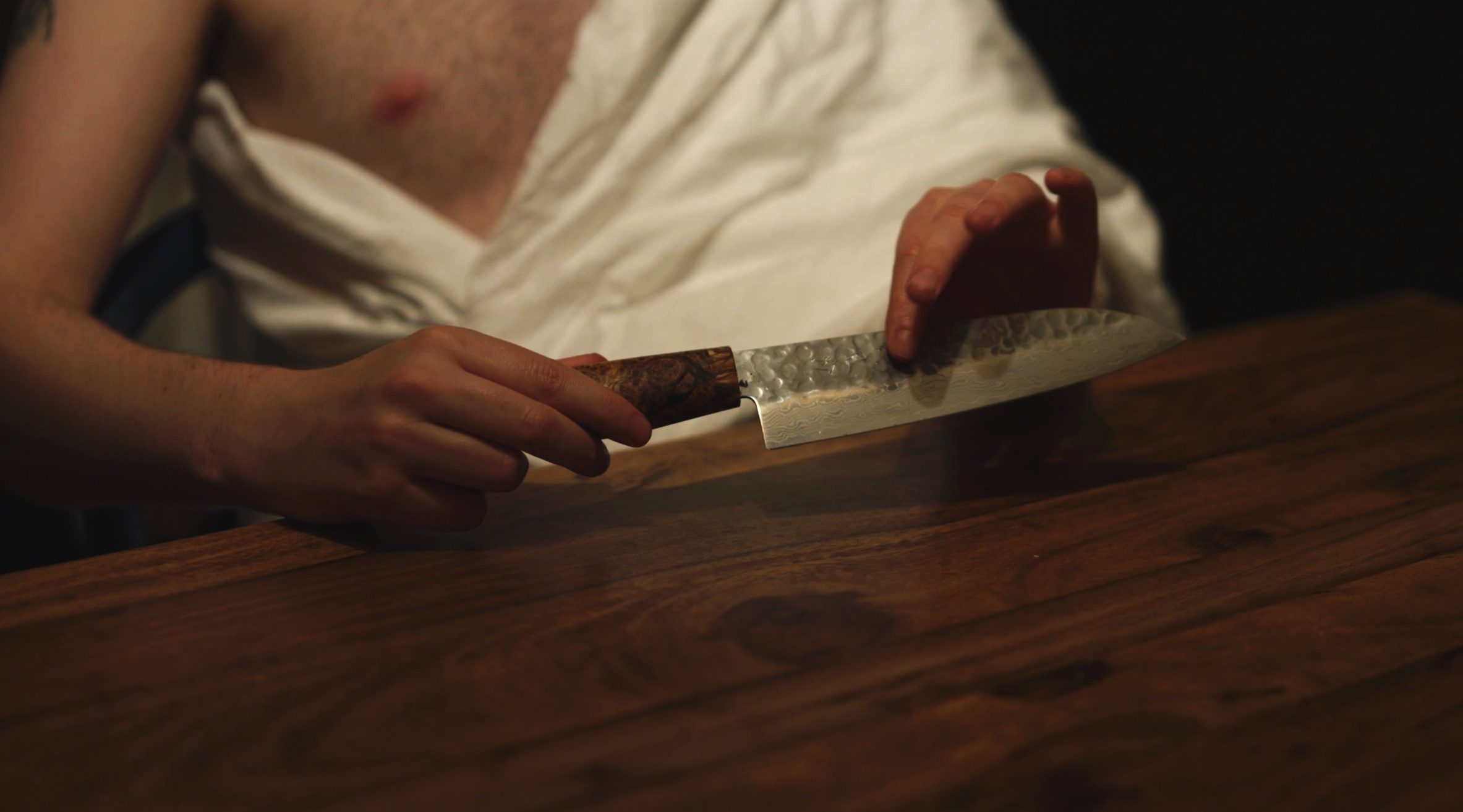 Person slicing something on a wooden surface with a large, textured knife, wearing a white apron.