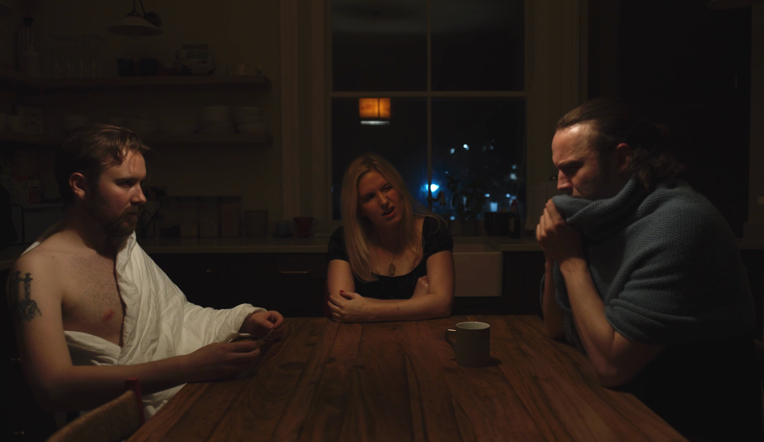Three people sitting at a wooden dinner table in a dimly lit kitchen at night. The woman in the middle has blonde hair and is wearing a black top, and looks concerned. The man on the left, with a beard and short hair, is shirtless with a white shirt 