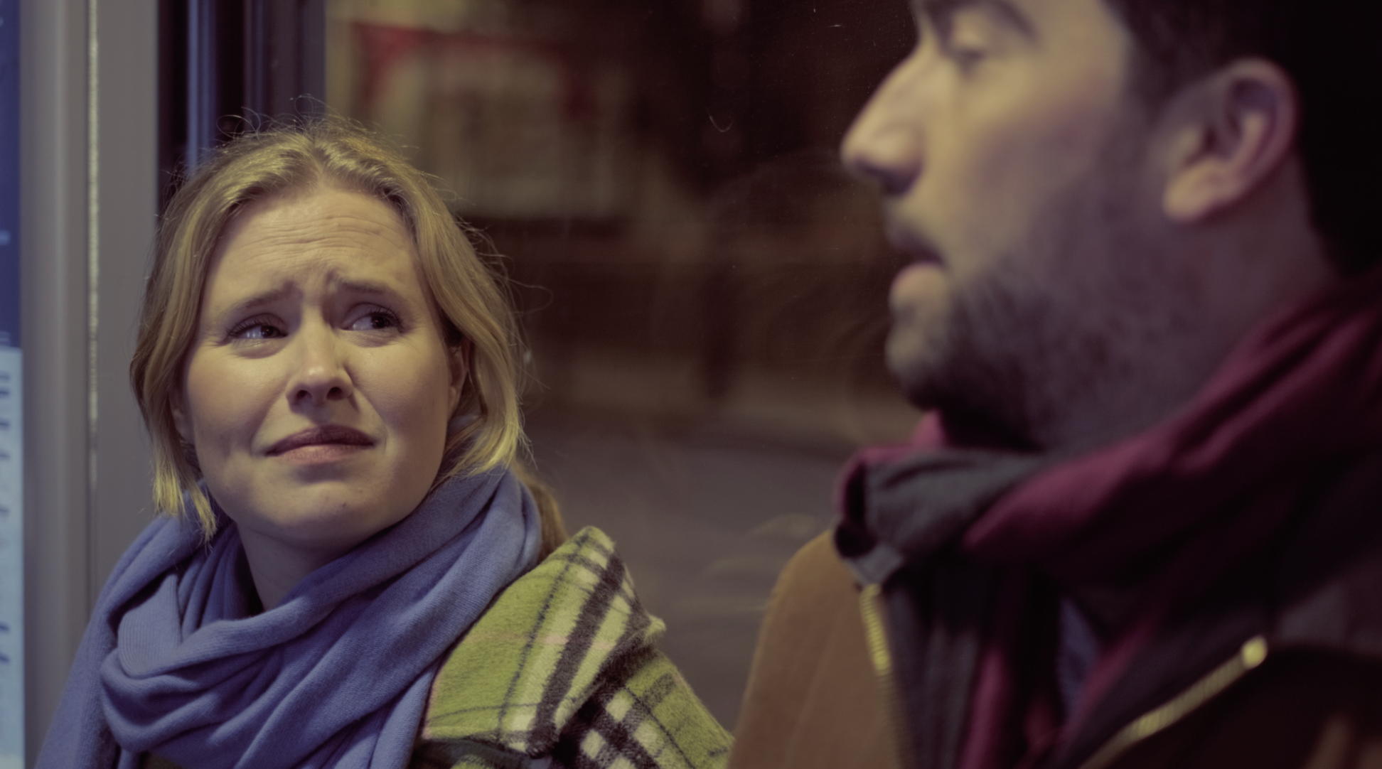 A woman with blond hair and a lavender scarf looks concerned while talking to a man with dark hair and a beard, wearing a maroon jacket and in a hooded hoodie inside a public transportation vehicle.