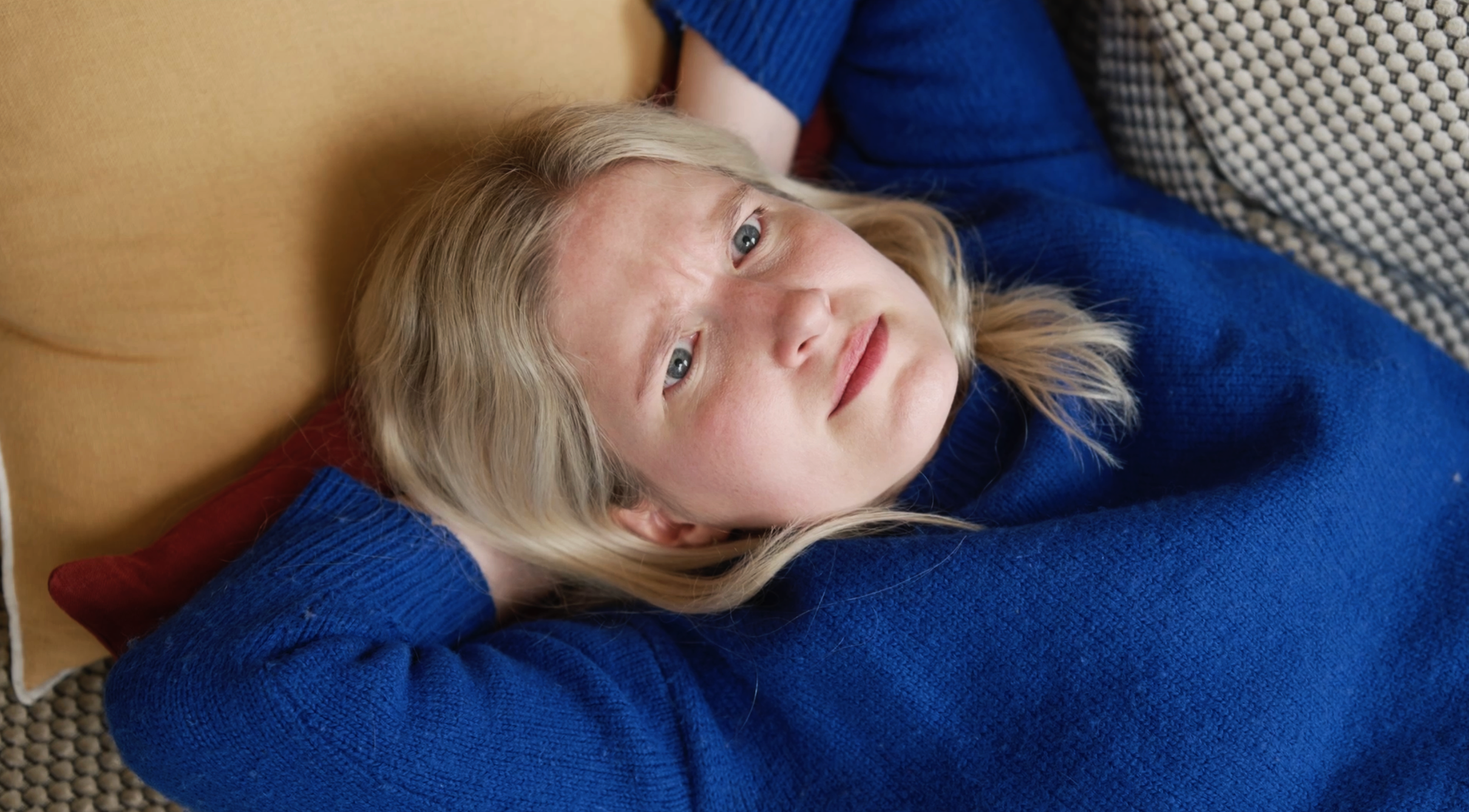A woman with blonde hair lying on a bed, wearing a blue sweater, looking thoughtfully upwards with her arms behind her head.