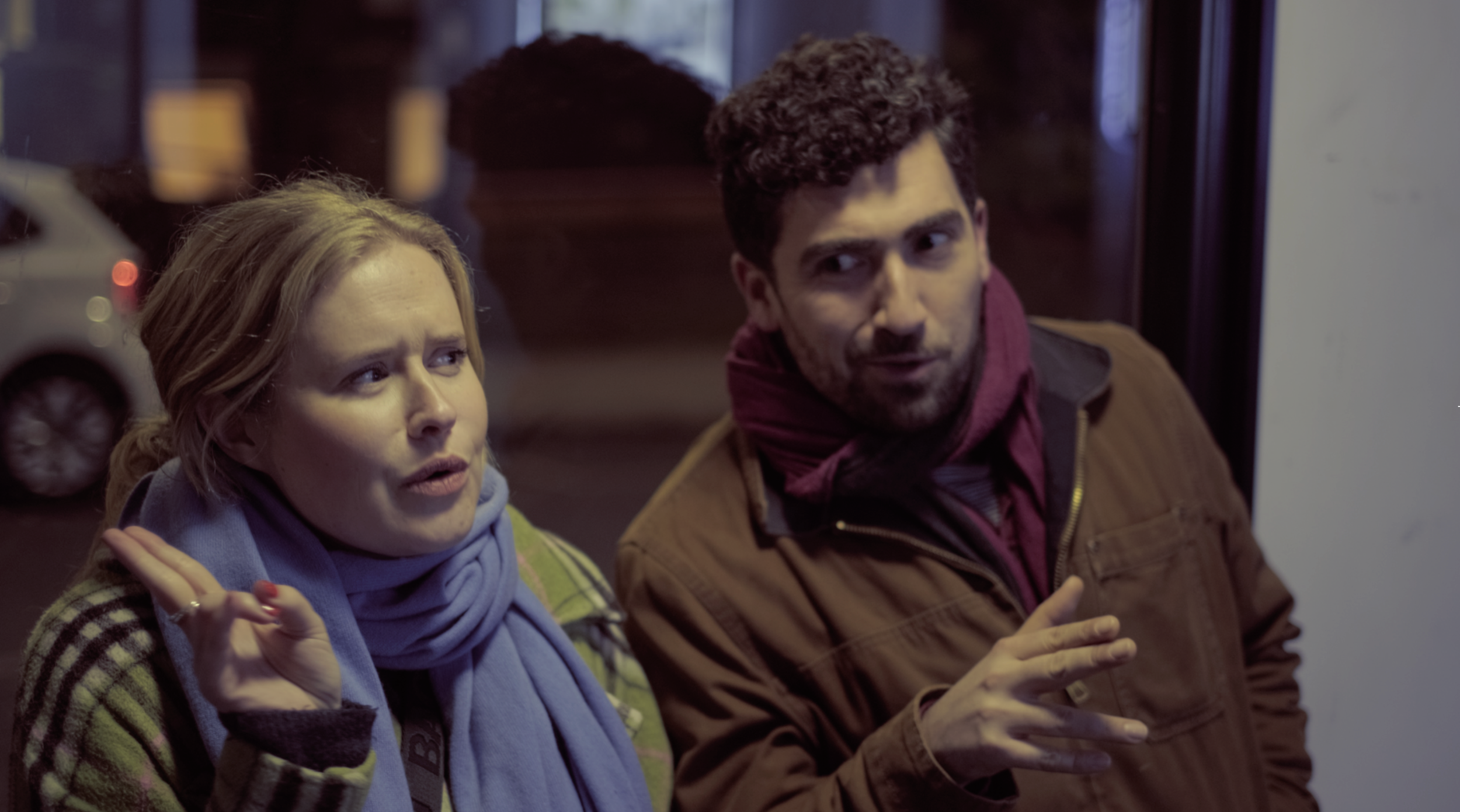 Two people, a woman and a man, are engaged in a conversation outside at night. The woman has blonde hair, wears a checkered coat and a blue scarf, and appears to be speaking. The man has dark hair, a beard, and wears a brown jacket with a maroon scar