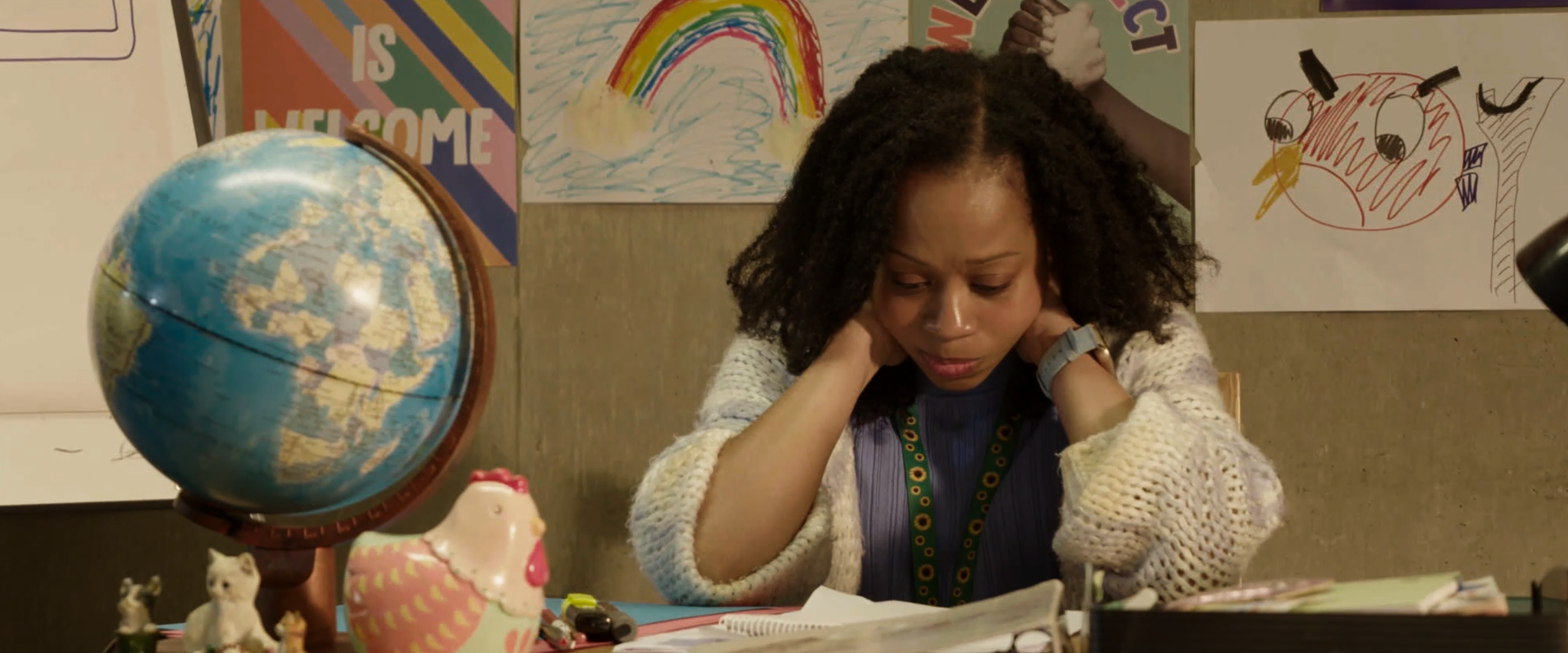 A woman sits at a cluttered desk, looking stressed or frustrated, with her hands on her neck. The background has children's drawings, including a rainbow and a bird. A globe, a pig figurine, a small animal figurine, and some stationery item are visib