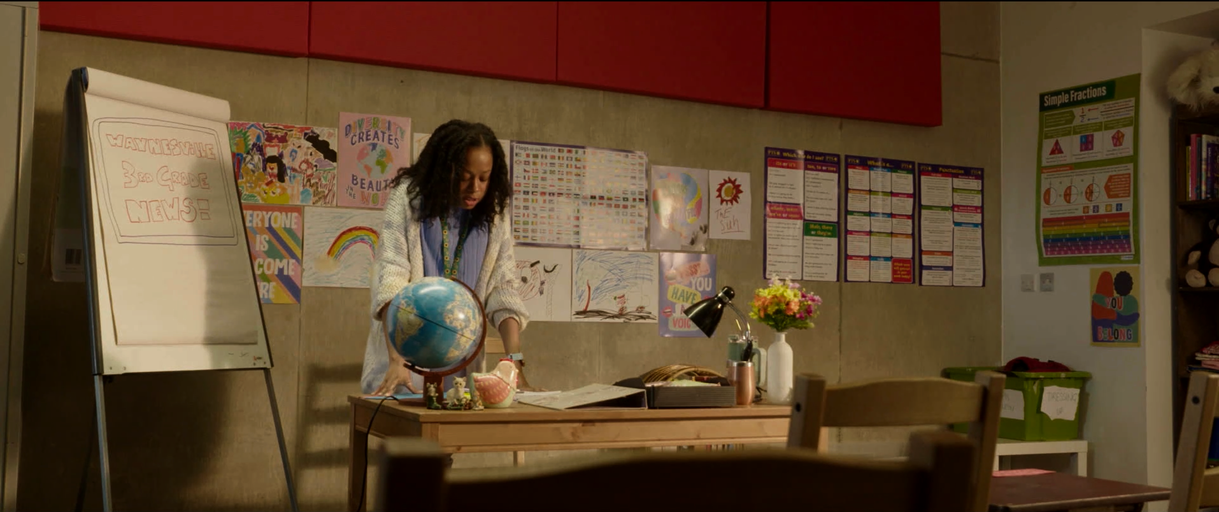 A woman organizing papers on a classroom desk with a globe, flower vase, and desk lamp, in front of a bulletin board decorated with children's artwork and educational posters.