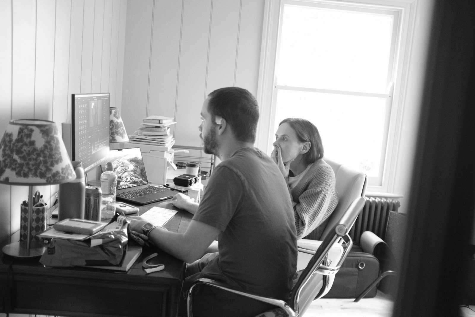 A man working at a desk with a woman watching behind him. The room has a window, a desk cluttered with papers, a laptop, and a monitor. The environment appears casual and home-like, in black and white.