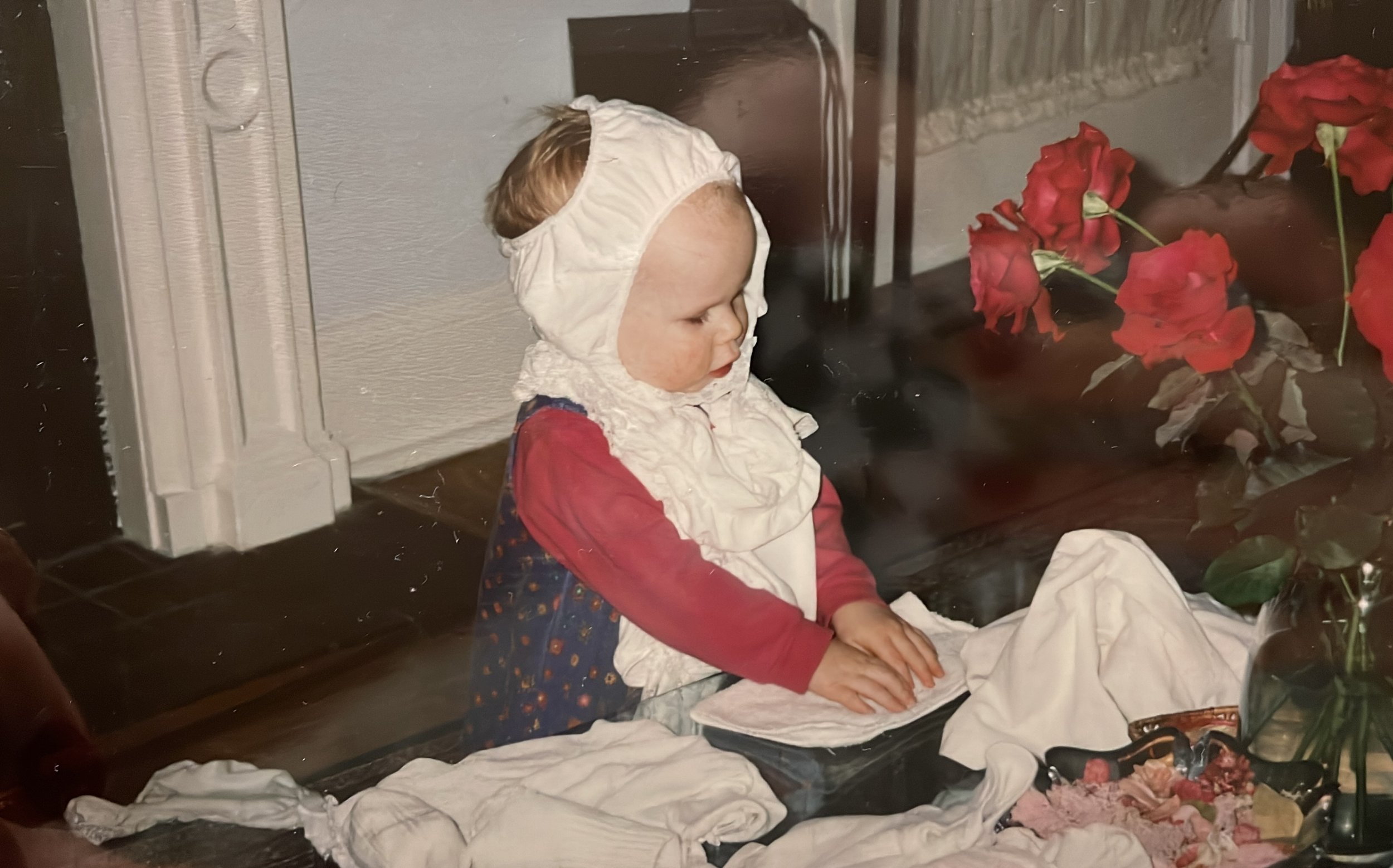 A young girl dressed in old-fashioned clothing, including a bonnet, sitting at a table with a spread of cloth and flowers nearby, appearing to be engaged in some activity.