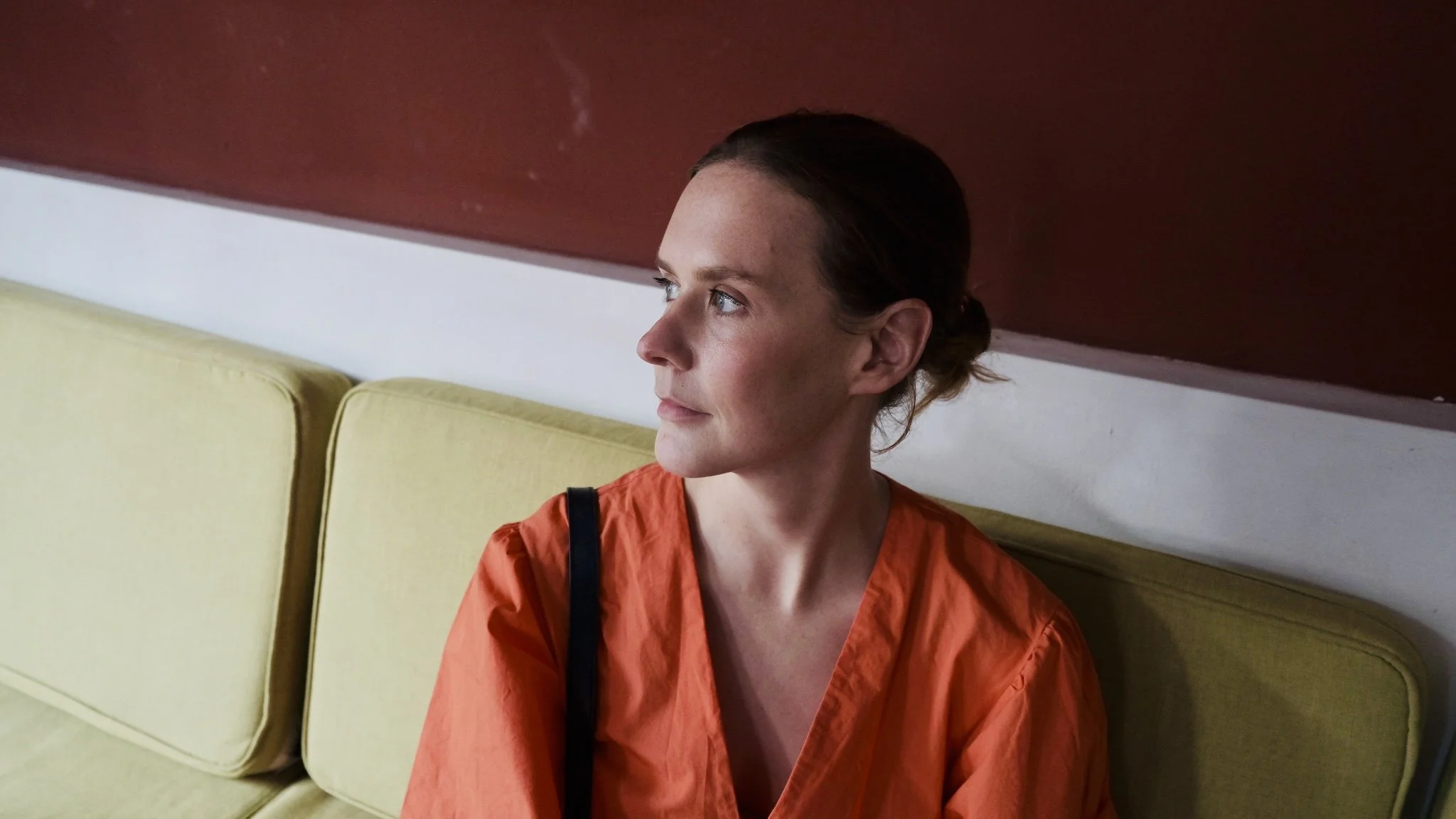 A woman with light skin and brown hair tied back, wearing an orange shirt, sitting on a yellow cushioned bench against a wall with a white stripe and a red section above, looking to her left.