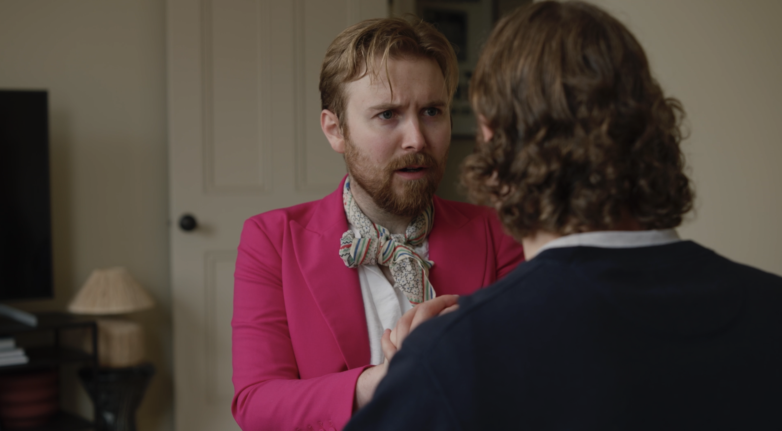 A man with a surprised or confused expression is facing a person with curly hair, holding their hands. The scene is set in a room with a closed door and a lamp in the background.