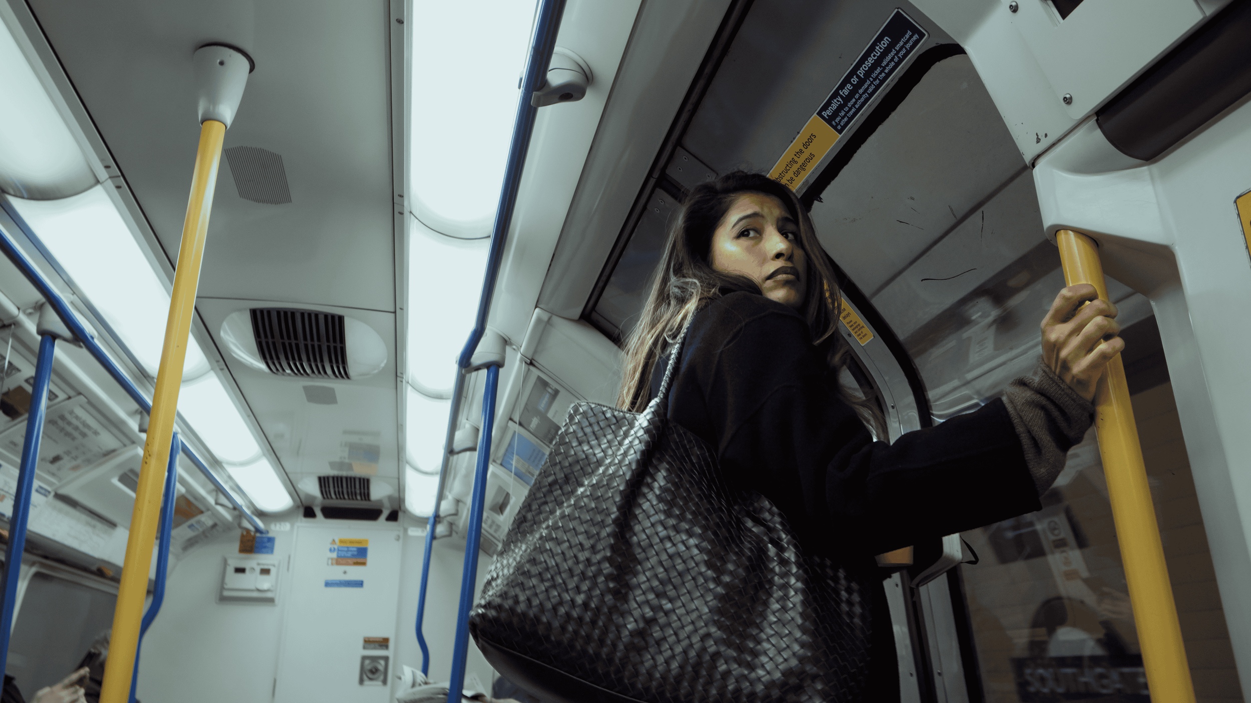 A young woman with long hair and a black jacket, carrying a patterned bag, holding onto a yellow pole inside a bus, looking over her shoulder.