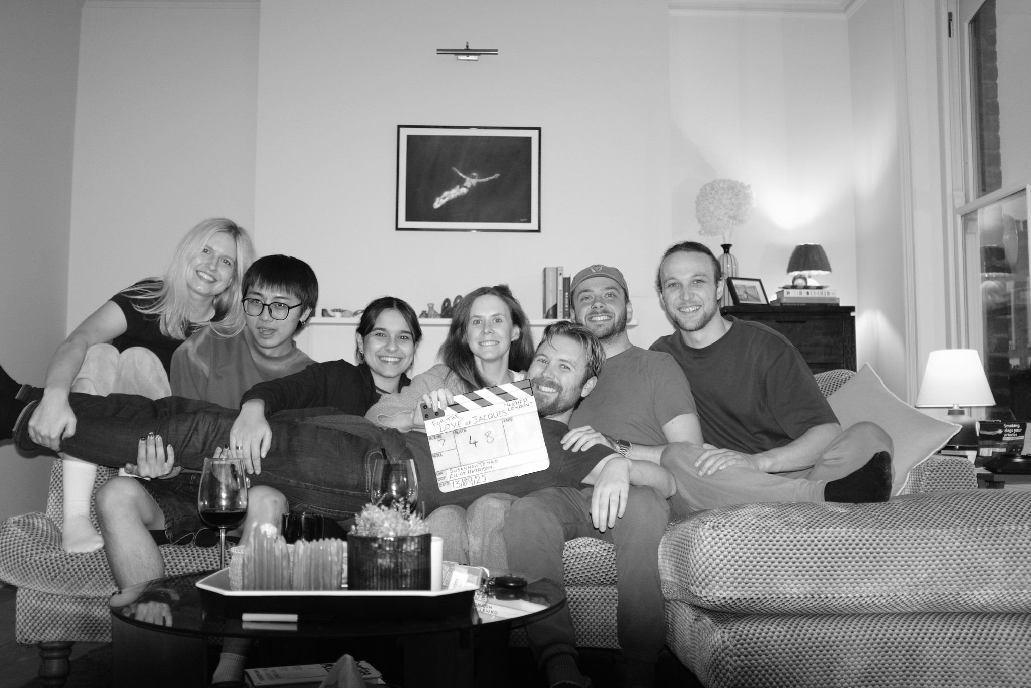 A group of seven people sitting and lying on a couch in a living room, smiling at the camera during a film or photo shoot, with a clapperboard in the center, wine glasses, and a coffee table with snacks and drinks in front of them.