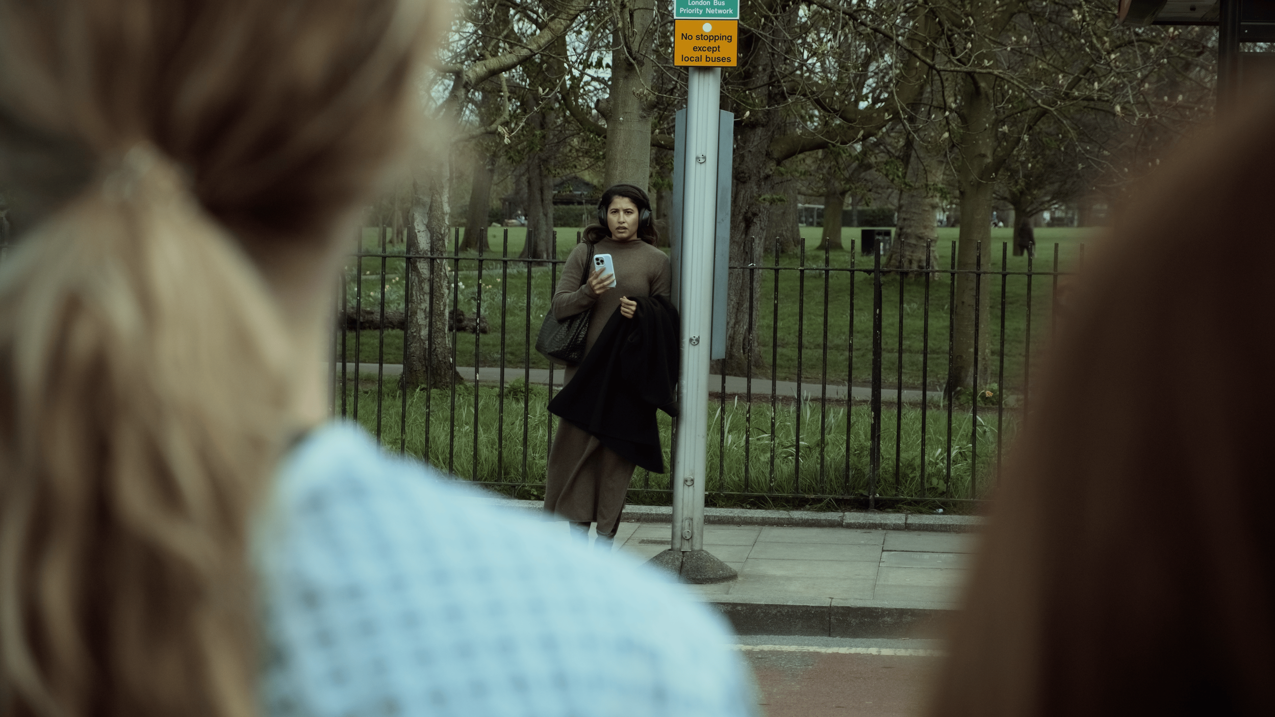 A woman stands at a bus stop holding a phone, with a black coat draped over her arm, and wearing headphones. She is looking at the camera. Two other women are in the foreground, out of focus, with only parts of their heads visible. The scene is in a 