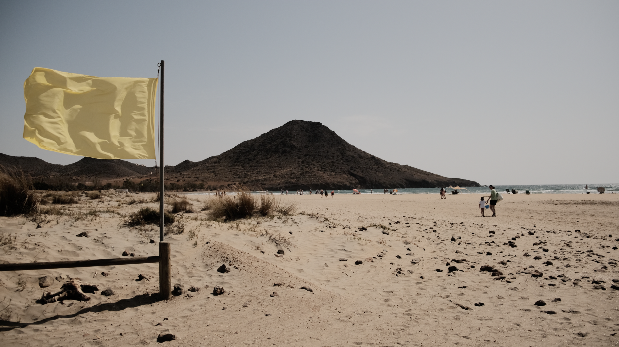 A deserted beach with a yellow flag on a pole, sandy shore, some patches of grass, and distant people near the water with a mountain in the background.