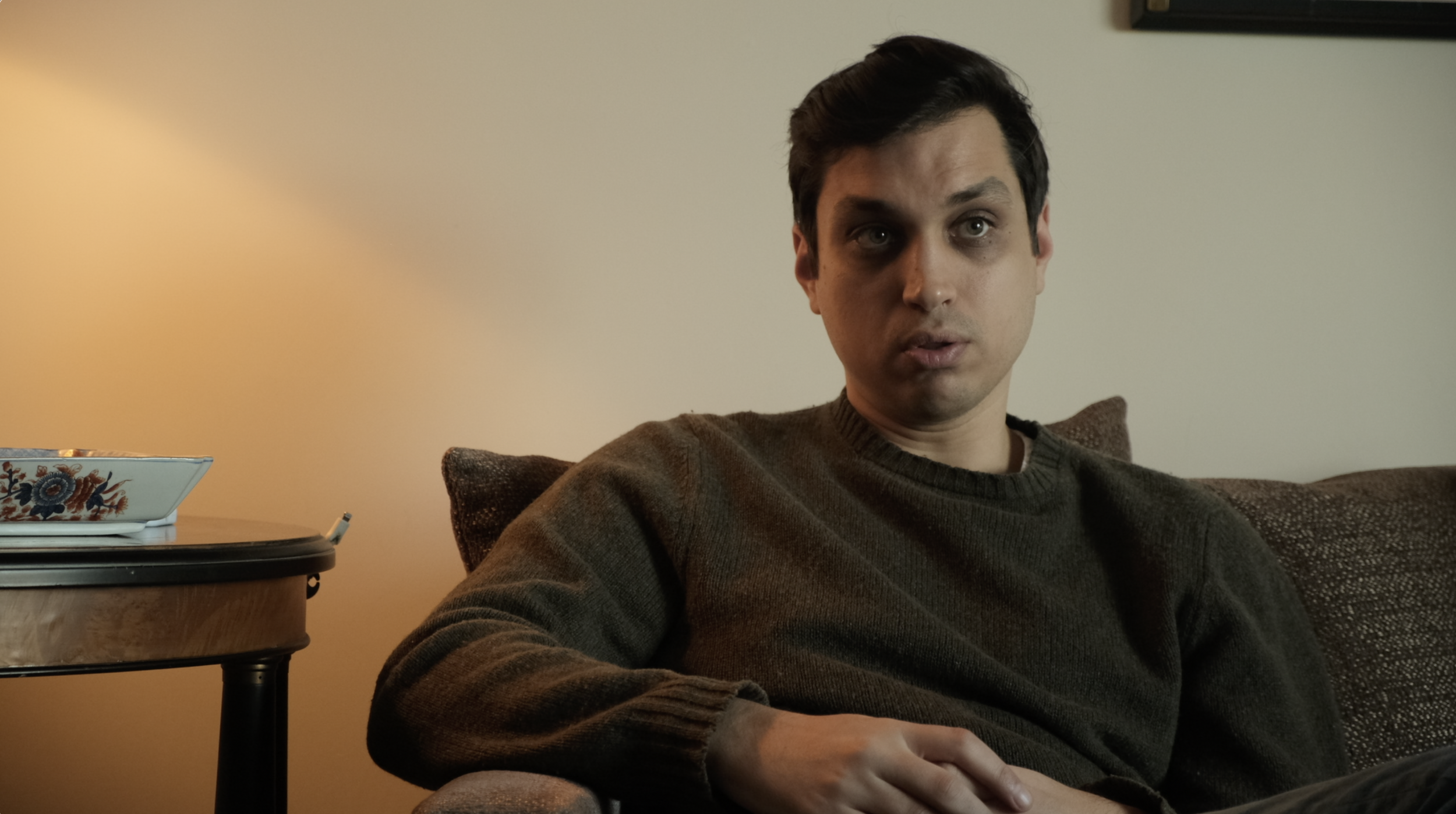A young man sitting on a couch with a brown sweater, looking slightly to the side with a neutral expression in a living room with a side table and a decorative bowl.