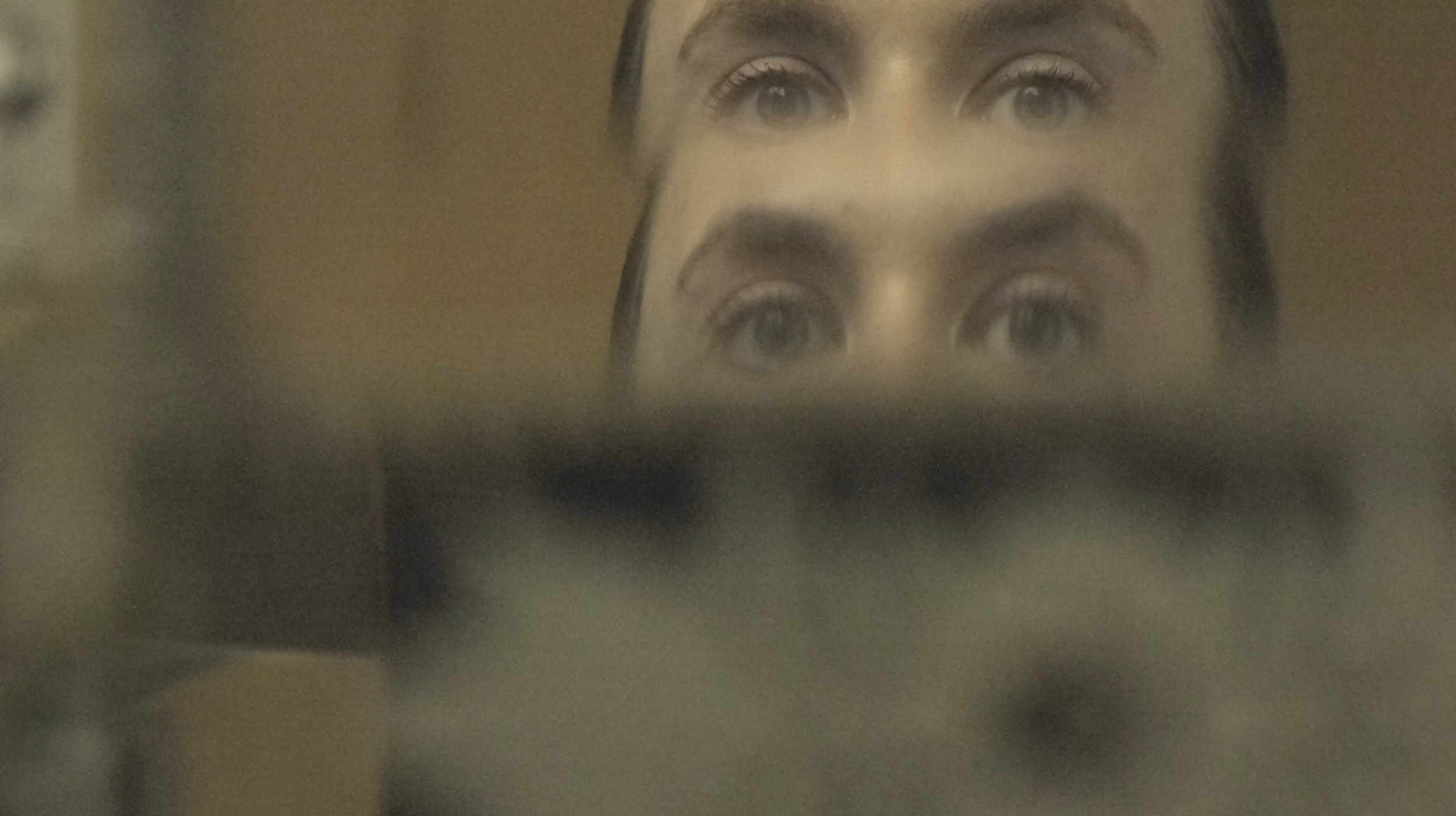 A reflection of a woman's face on a mirror, showing her eyes and forehead, with a blurry foreground object partially obscuring her face.