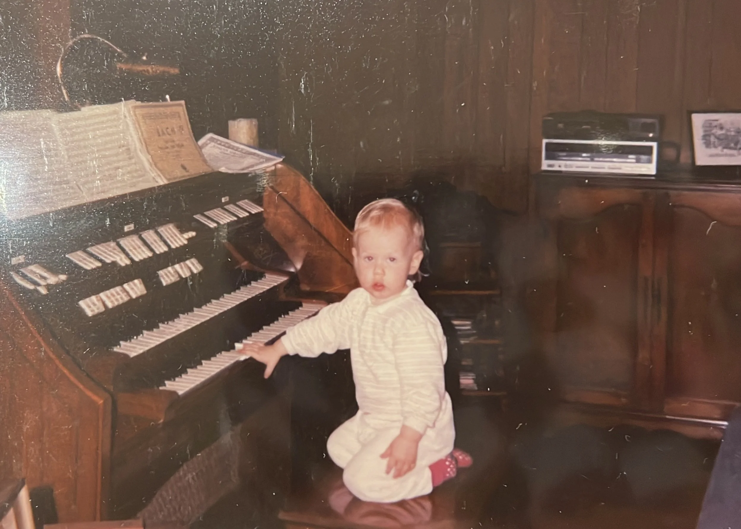A young child with blonde hair, wearing a light-colored striped outfit, sitting on a piano bench with one hand on the keys, in a room with wooden wall paneling and furniture, a stereo system on the right, and sheet music on an open piano.