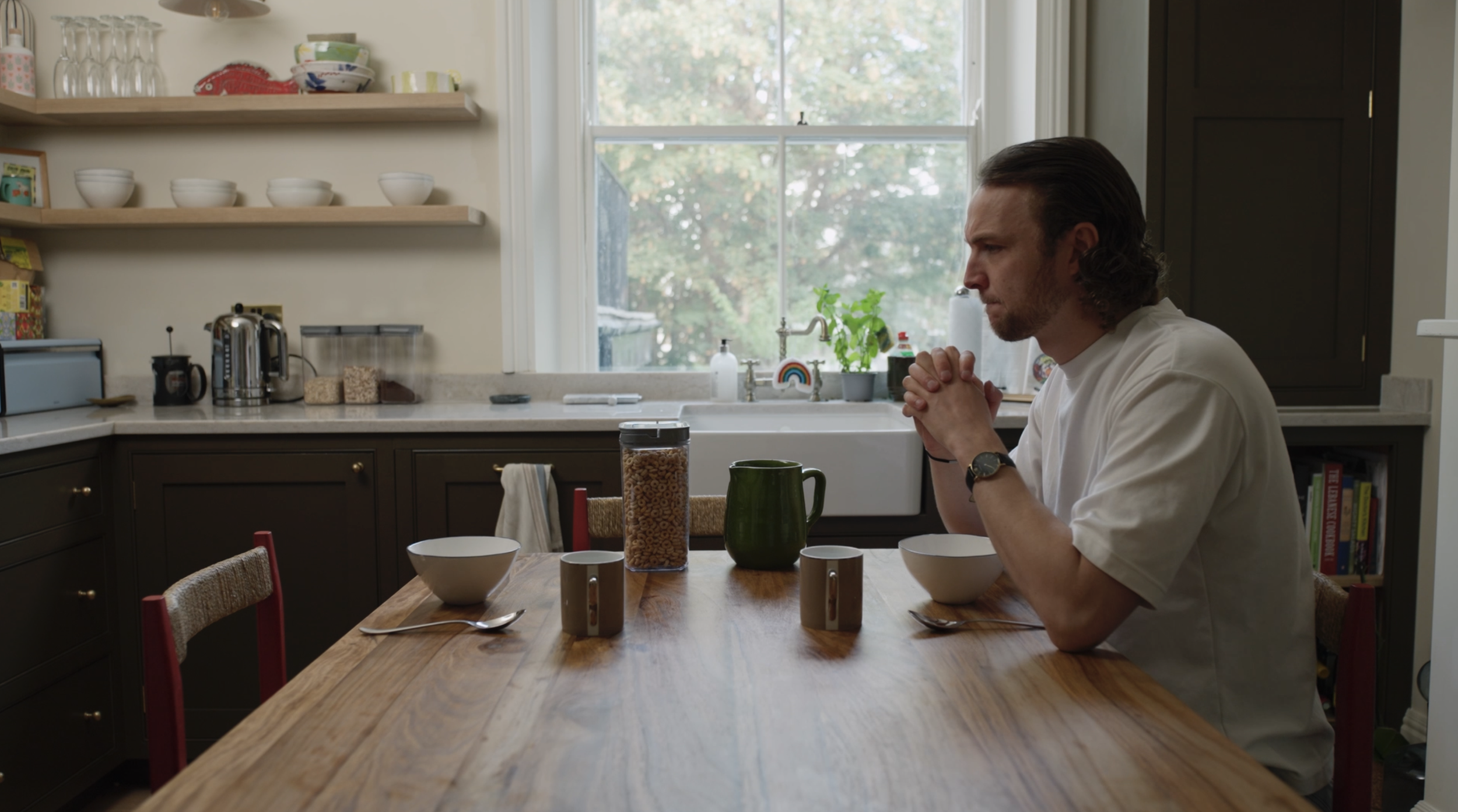 A man with long hair and a beard, wearing a white shirt, sits at a wooden dining table in a kitchen, praying with hands clasped. The table has two bowls, two mugs, a pitcher, and a container of cereal. The kitchen has open shelves with bowls, glasses