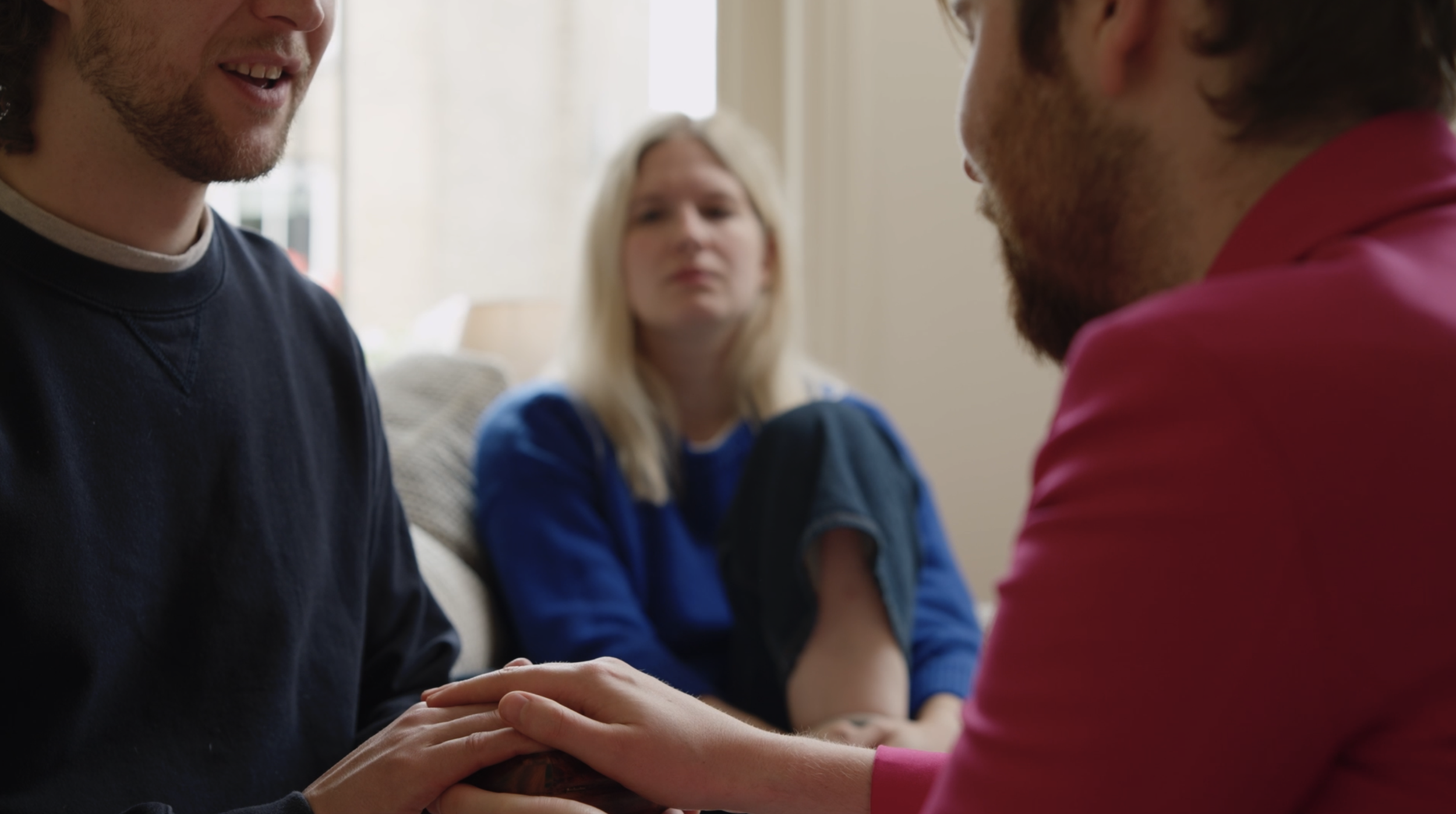 Two men holding hands and gazing at each other during a heartfelt moment, with a woman in the background watching with concern.