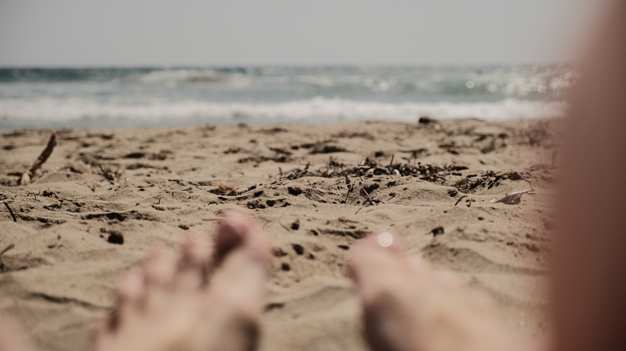 Close-up of sandy beach with the ocean and waves in the background, as seen from a low angle with out-of-focus hands or fingers in the foreground.