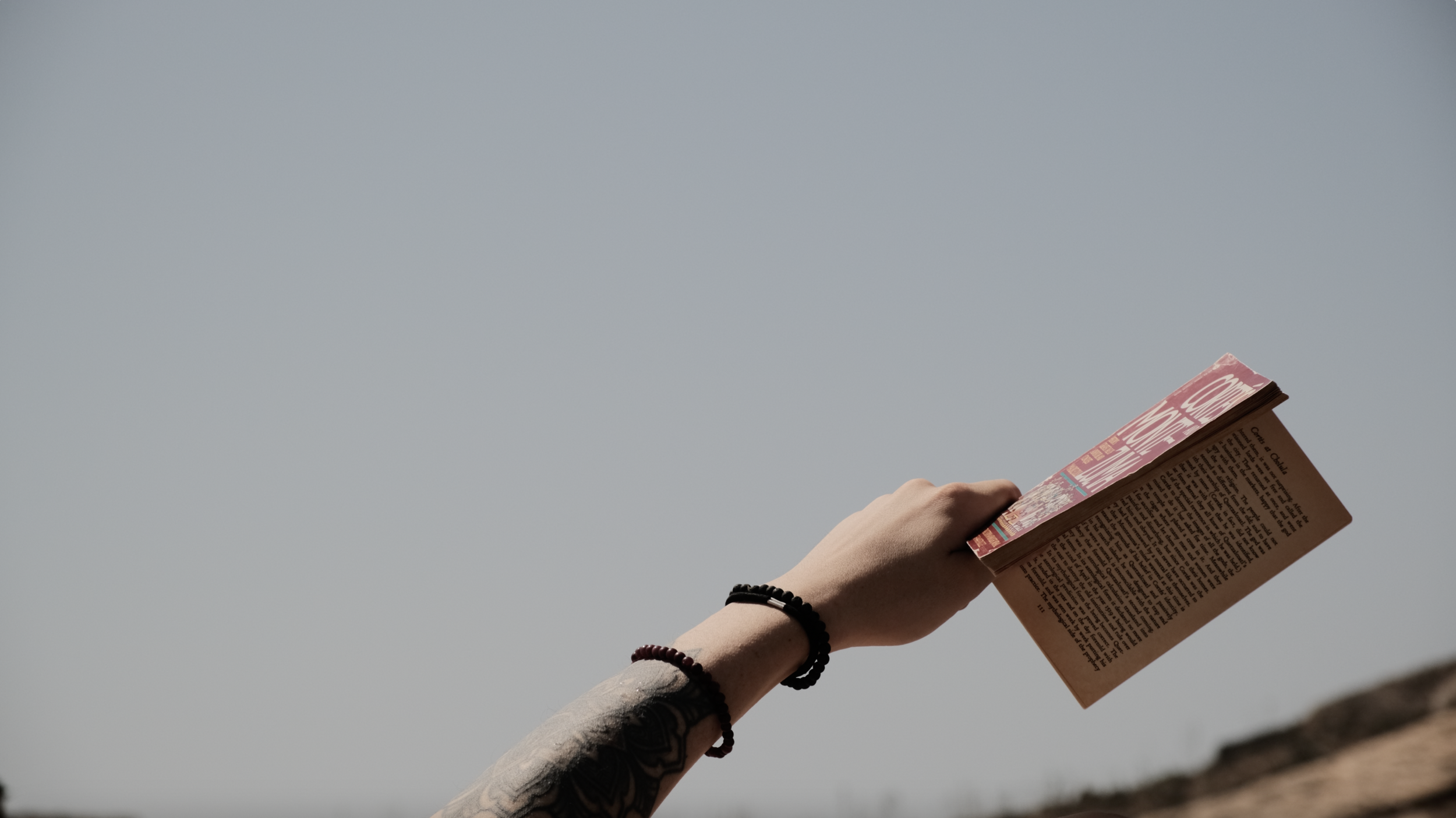 A person's arm holding a book against a clear sky, with a tattoo on the forearm and wearing two bracelets.