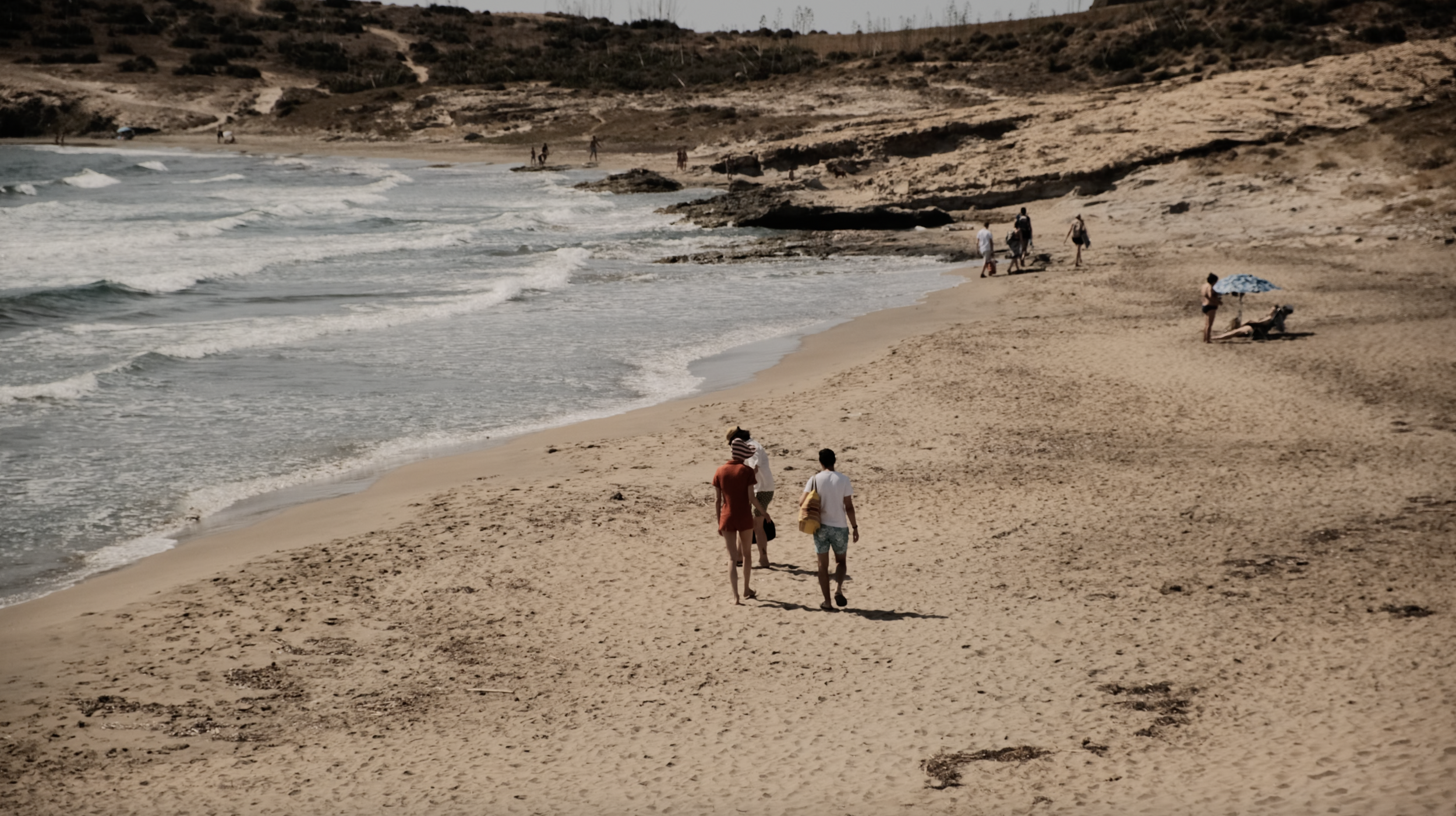 People walking along a sandy beach with some relaxing under umbrellas, and others near the shoreline with small waves.