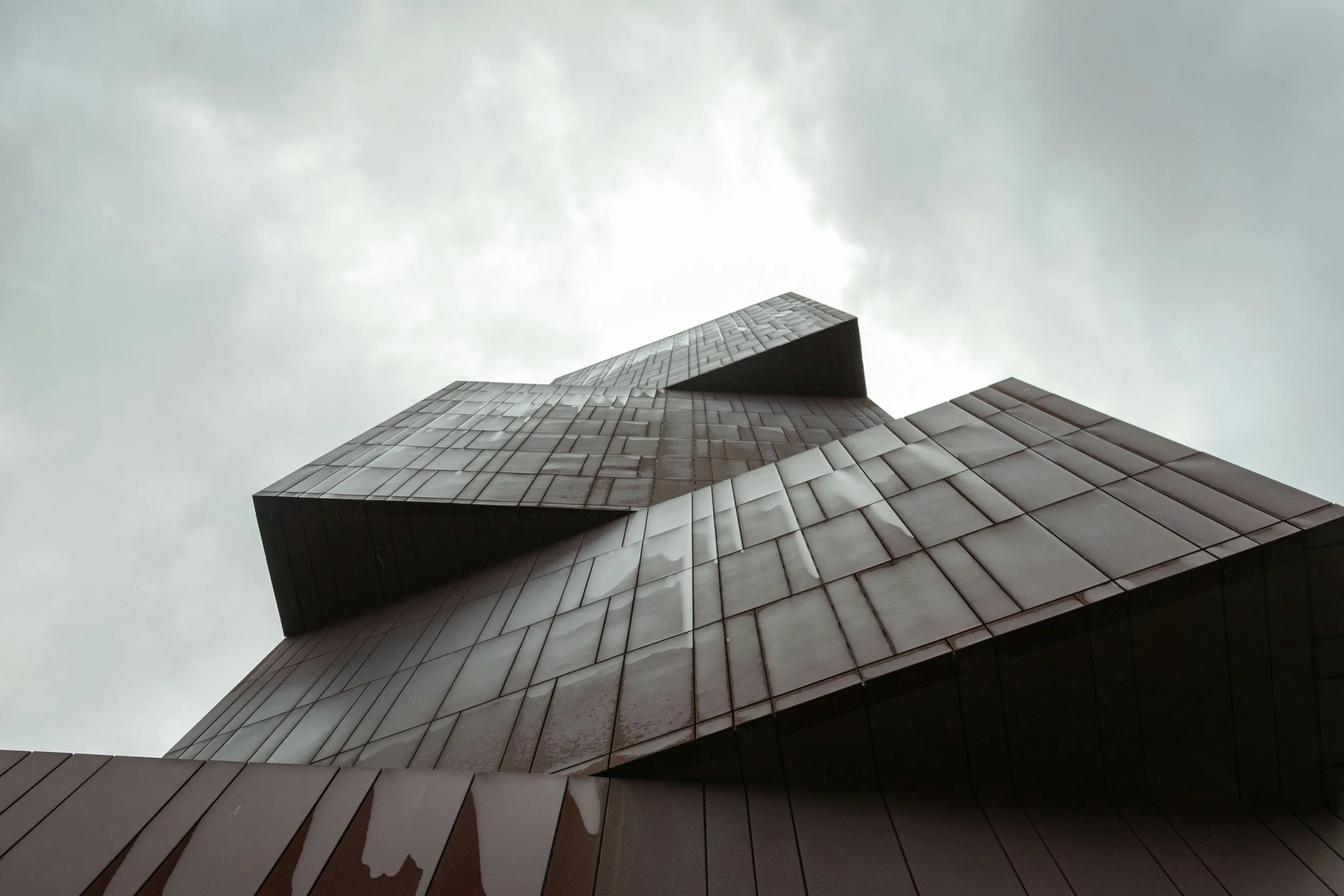 Looking up at a modern, angular building with reflective glass windows and a gray, cloudy sky.