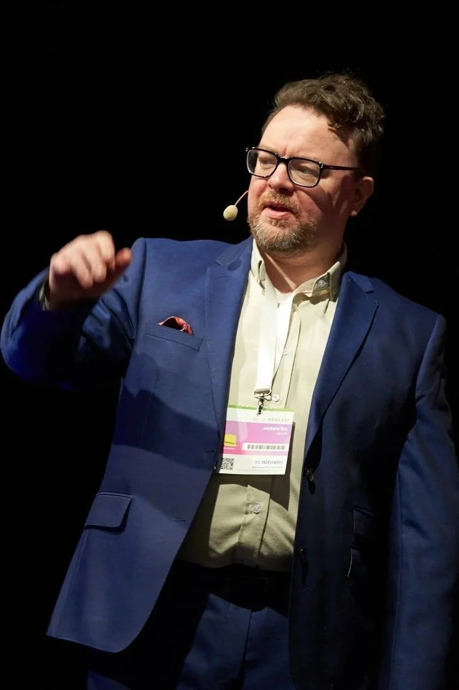 A man with glasses and a beard dressed in a blue suit and white shirt, speaking at a conference or event with a microphone headset.