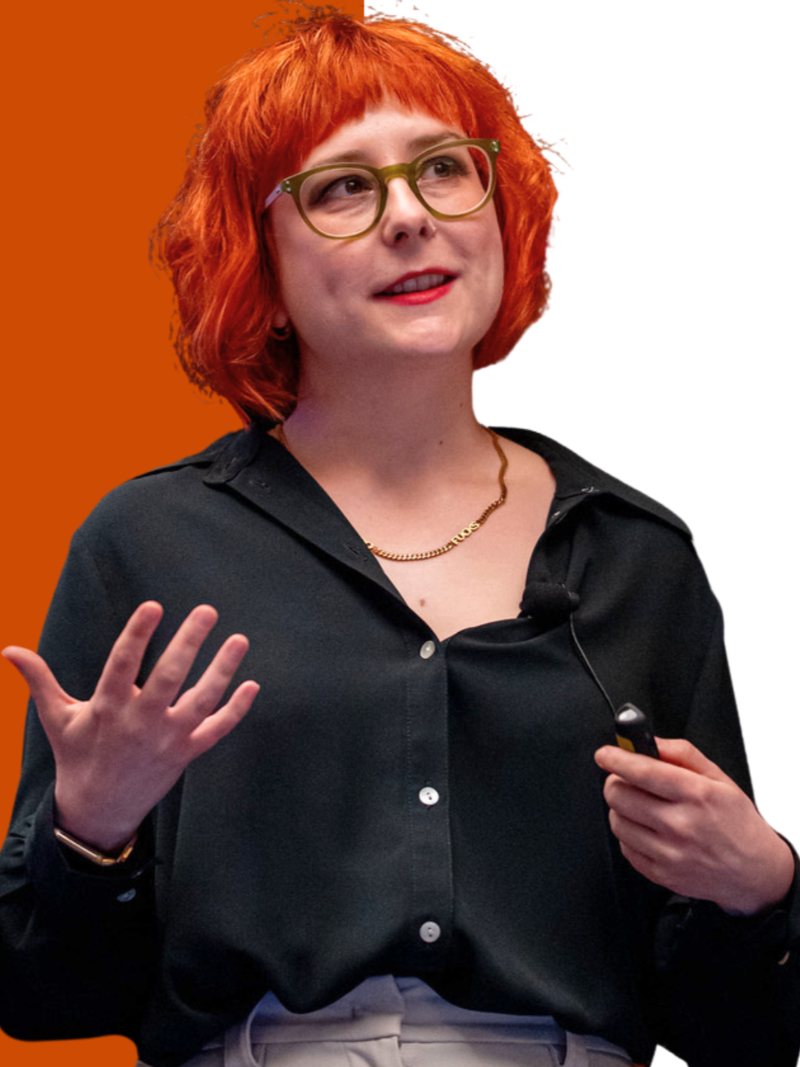 A woman with red hair, green glasses, and a black shirt is speaking and gesturing with her left hand, holding a remote in her right hand against a background that is partially orange and white.