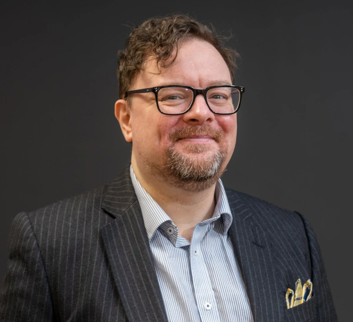 Portrait of a man with glasses, smiling, wearing a pinstripe suit and a patterned shirt, against a dark background.