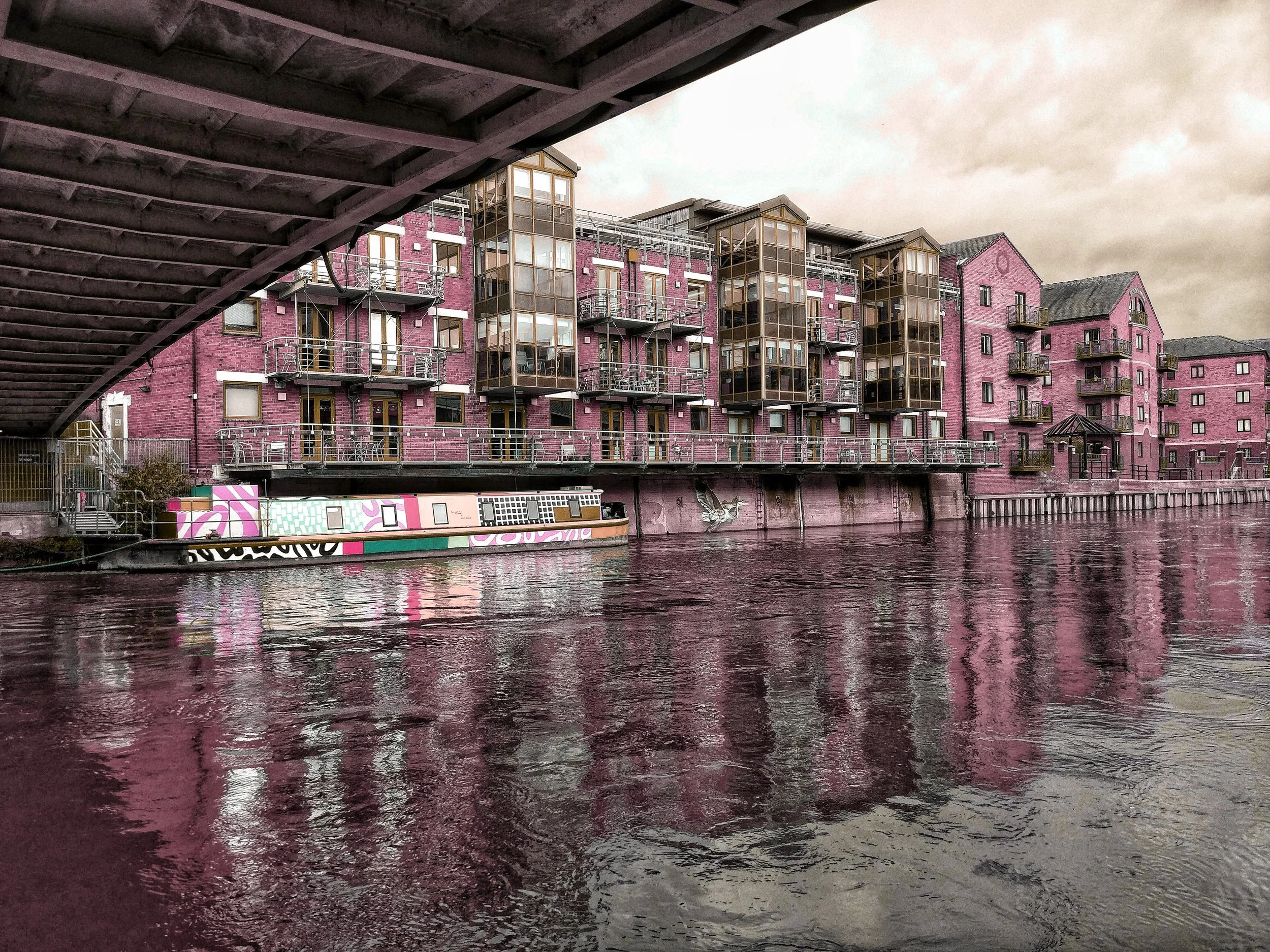 A large pink brick apartment complex along a river, with a bridge overhead and graffiti art on a boat docked along the water.