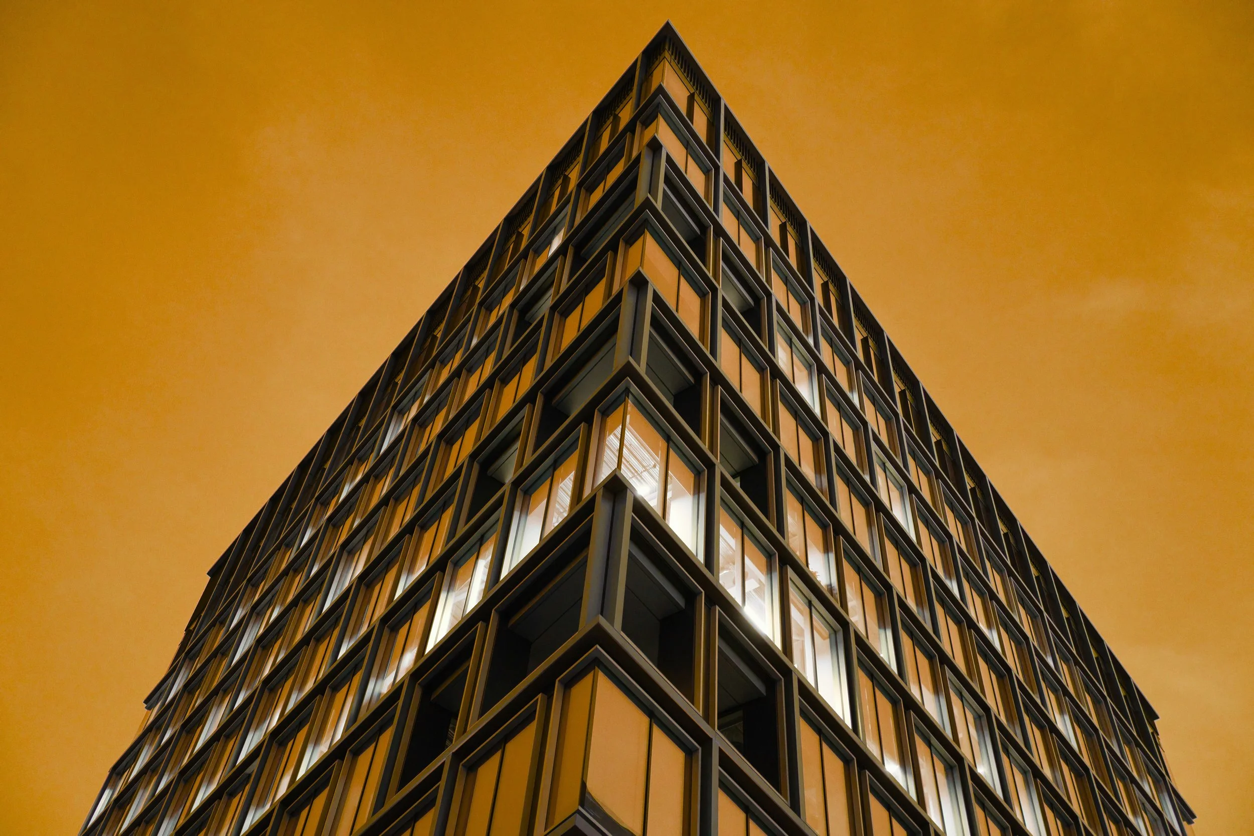 A modern multi-story building with illuminated windows against a yellow-orange evening sky.