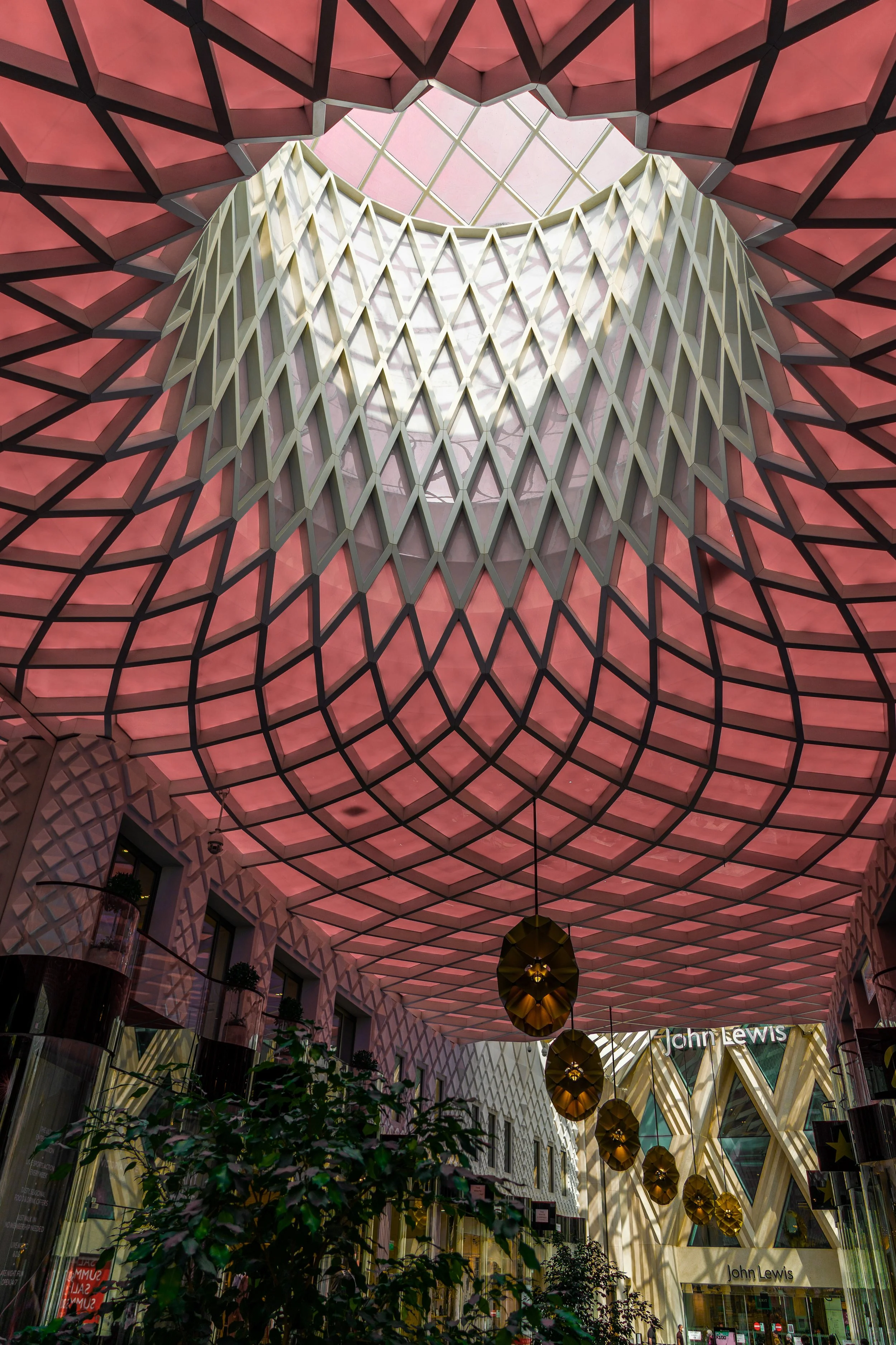 Interior view of a shopping mall with a large pink geometric mesh ceiling, hanging gold decorative lamps, and storefronts including a John Lewis store.