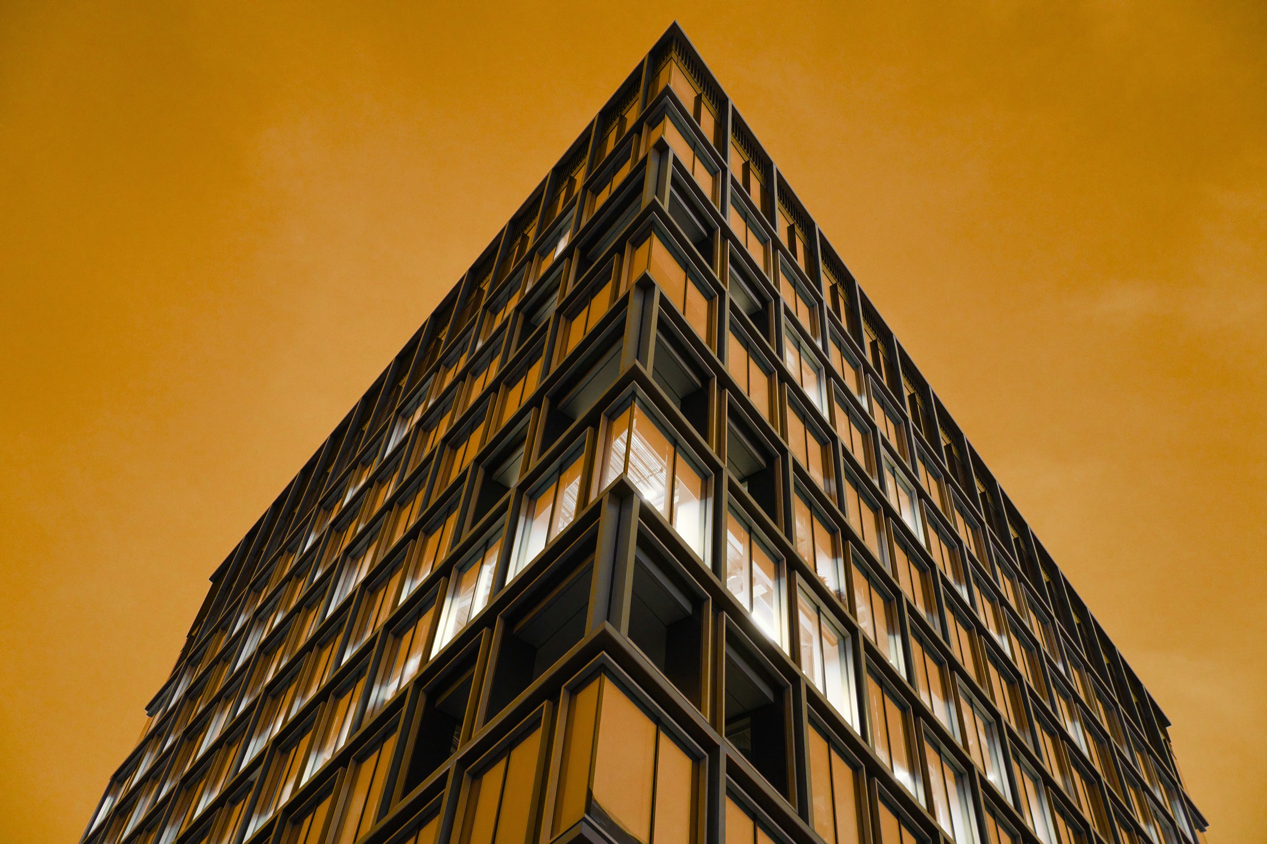 nighttime view of a modern, multi-story building with illuminated windows against an orange sky, taken from a low angle.
