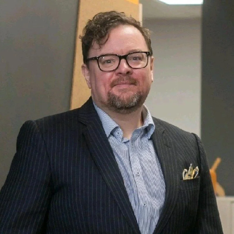 Man with glasses and a beard in a dark pinstripe suit, white shirt, standing indoors.