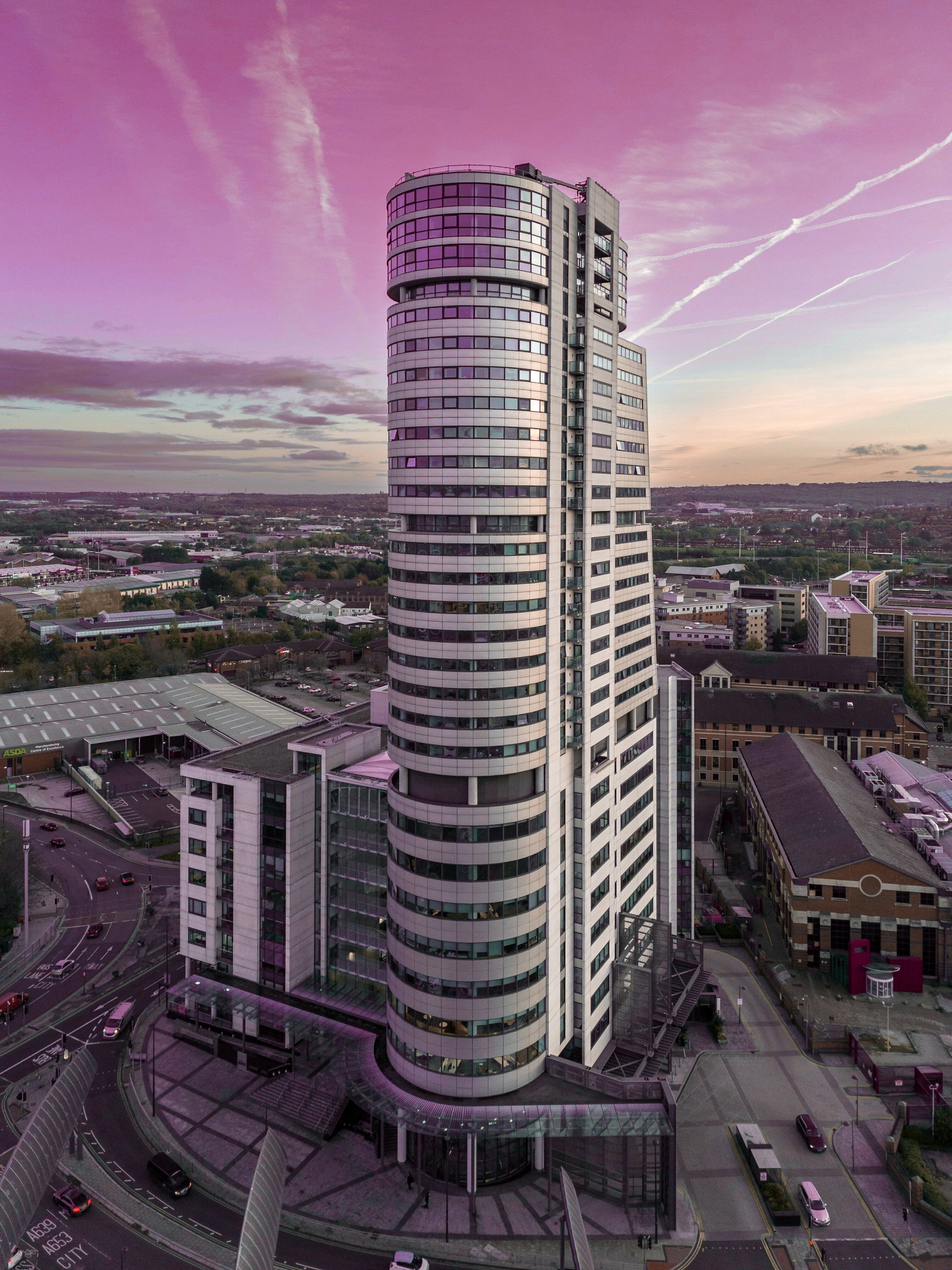A tall, modern skyscraper with a rounded top, reflecting a pink and purple sunset sky, surrounded by smaller buildings and streets.