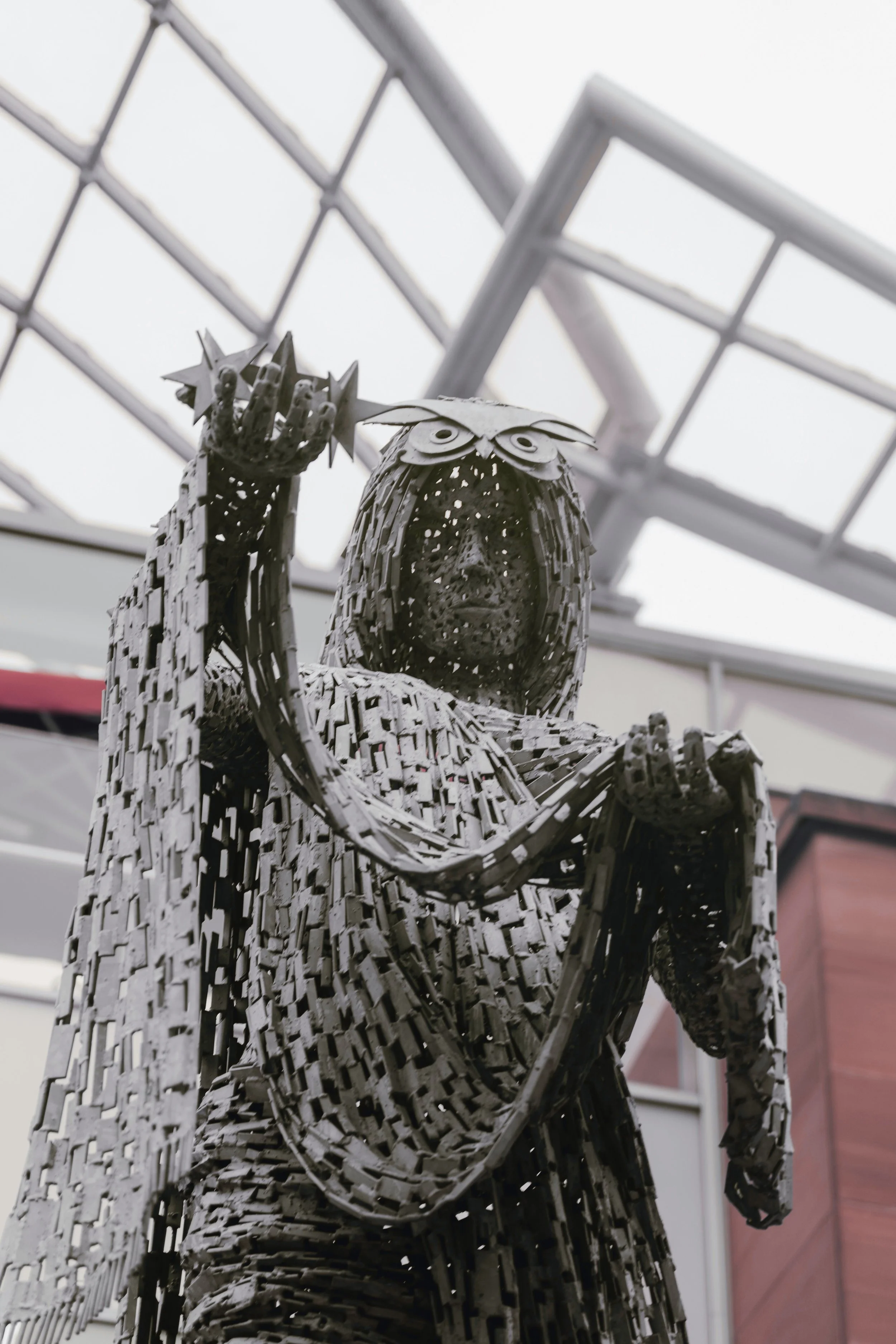 A metal sculpture of an owl wearing glasses, holding a star shaped object in its hand, located inside a building with a glass ceiling.