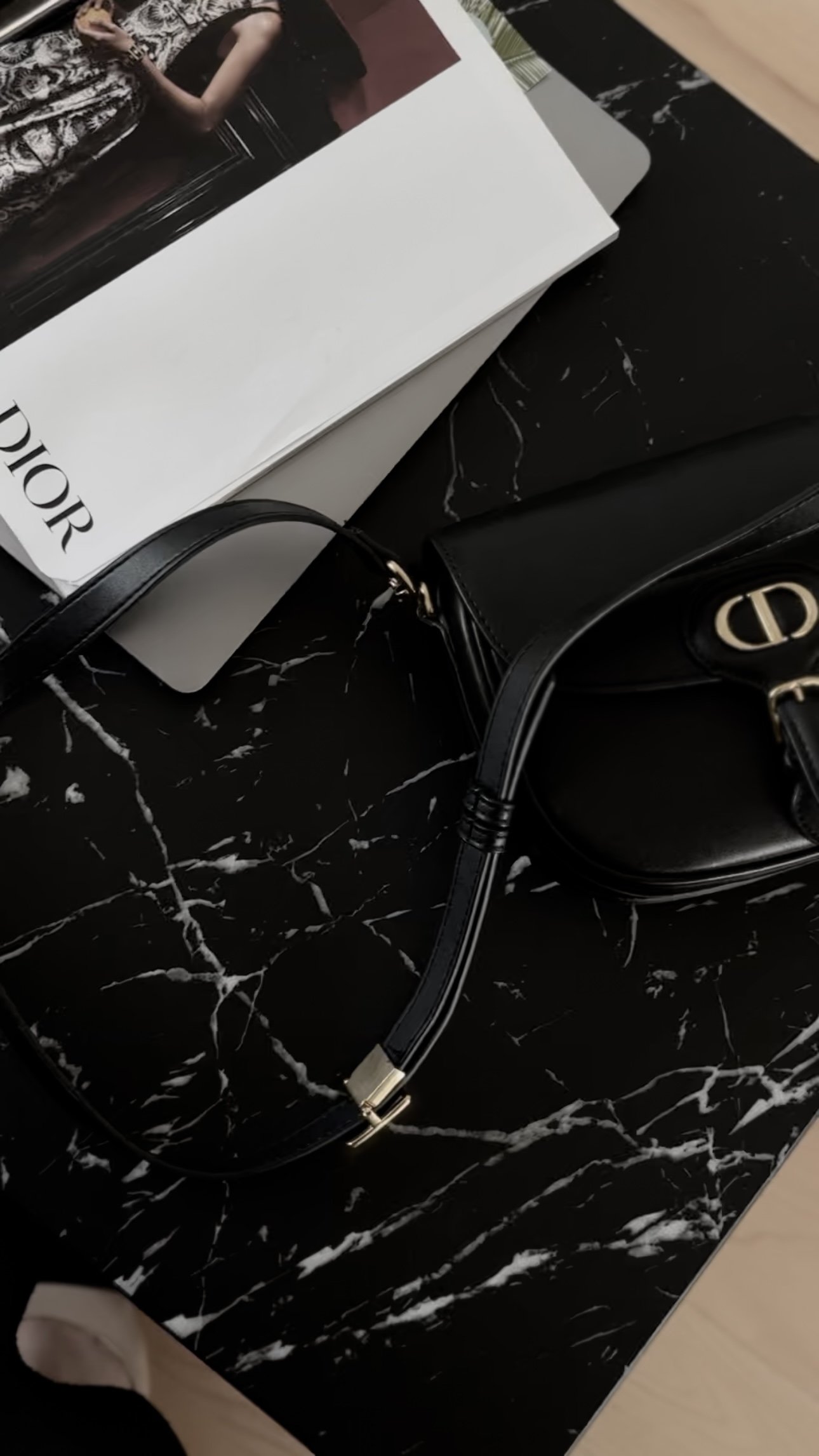 A close-up of a black Michael Kors handbag on a black marble surface. There is a Dior box in the background with a woman’s photo on it.