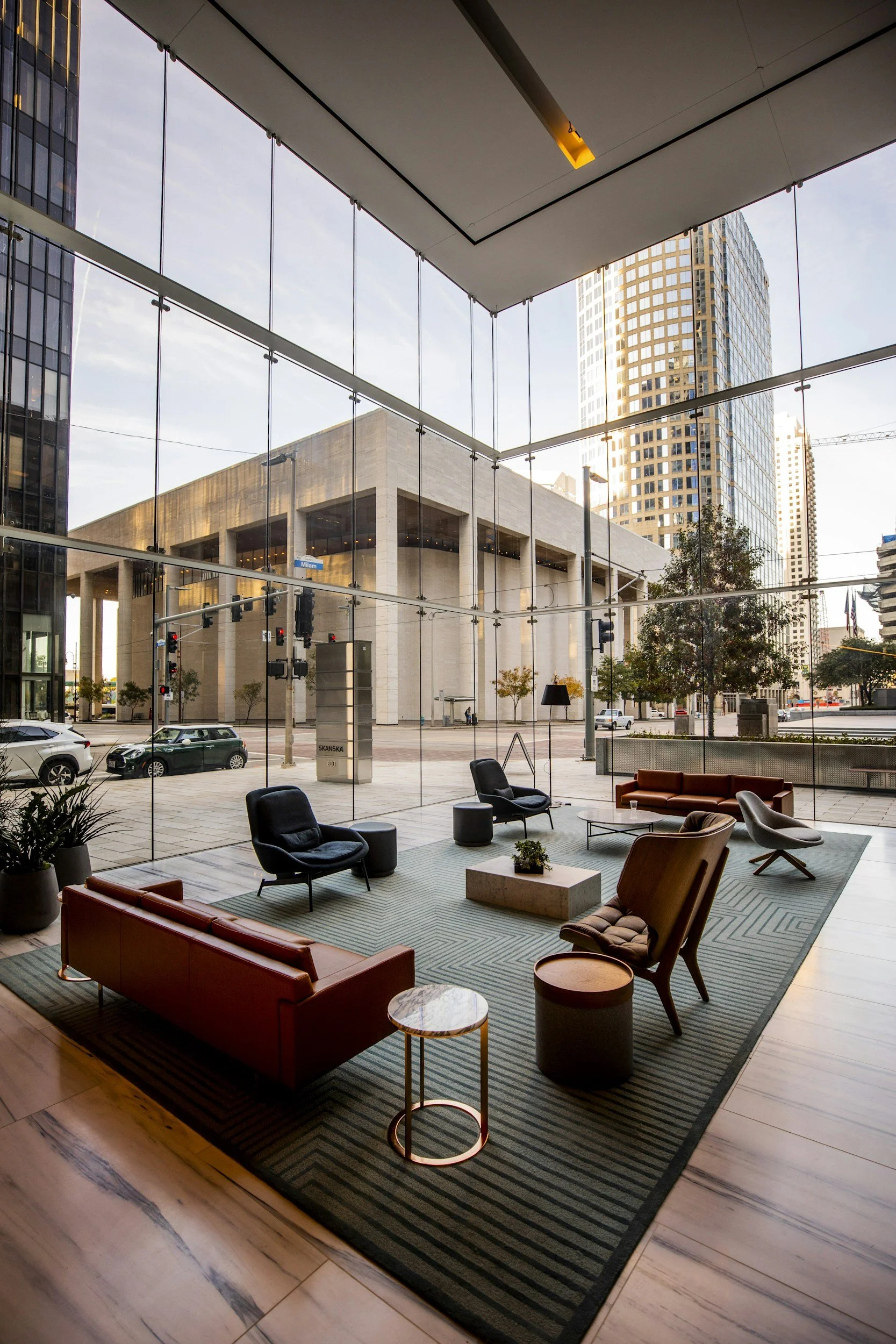 Modern hotel lobby with large glass windows, overlooking city streets and tall buildings, furnished with black and brown sofas, chairs, small tables, and a green rug.