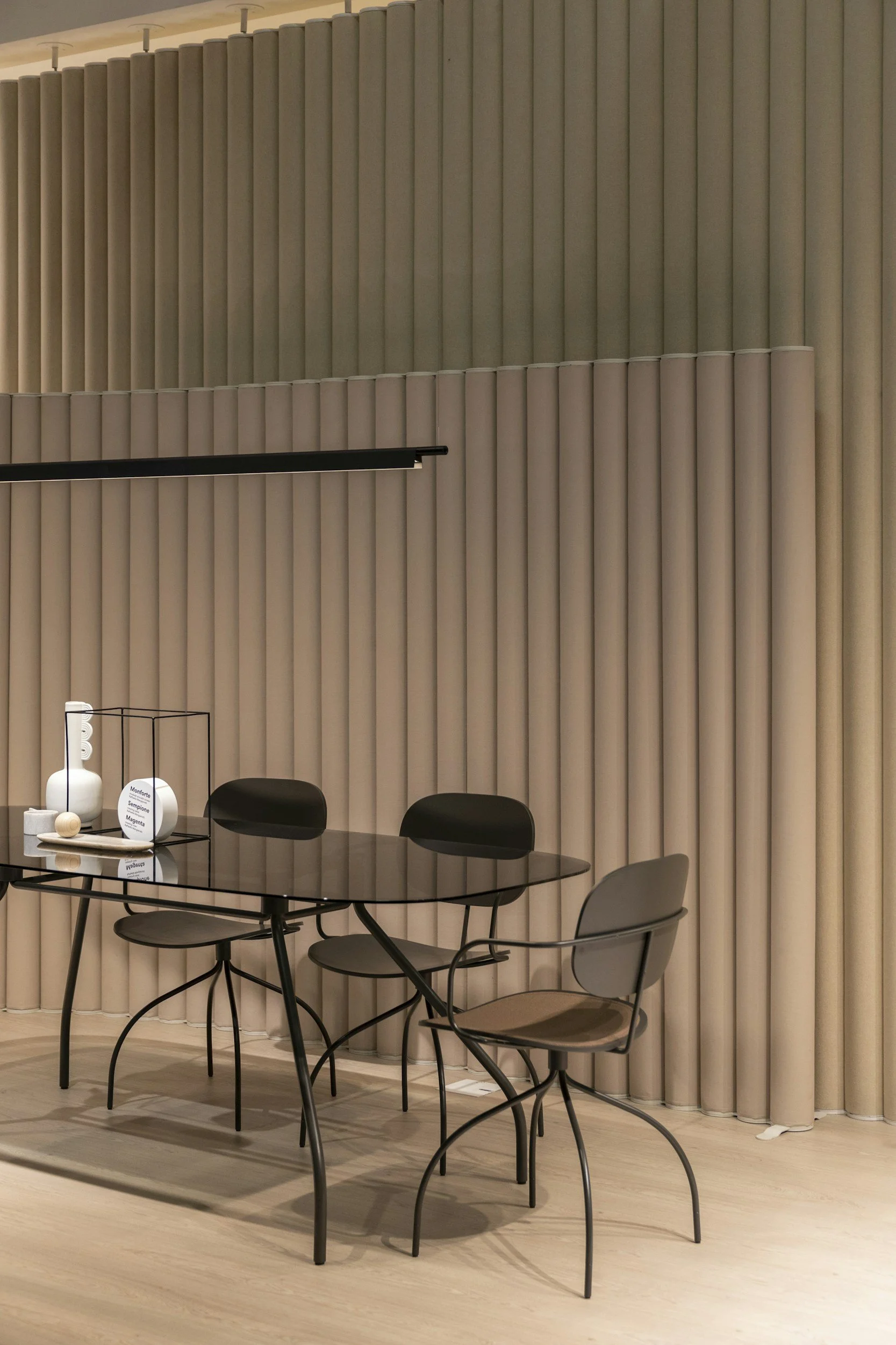 An interior space with beige vertical blinds on the wall, a black table with a minimalistic decoration on top, and four chairs around the table, one gray and three black.