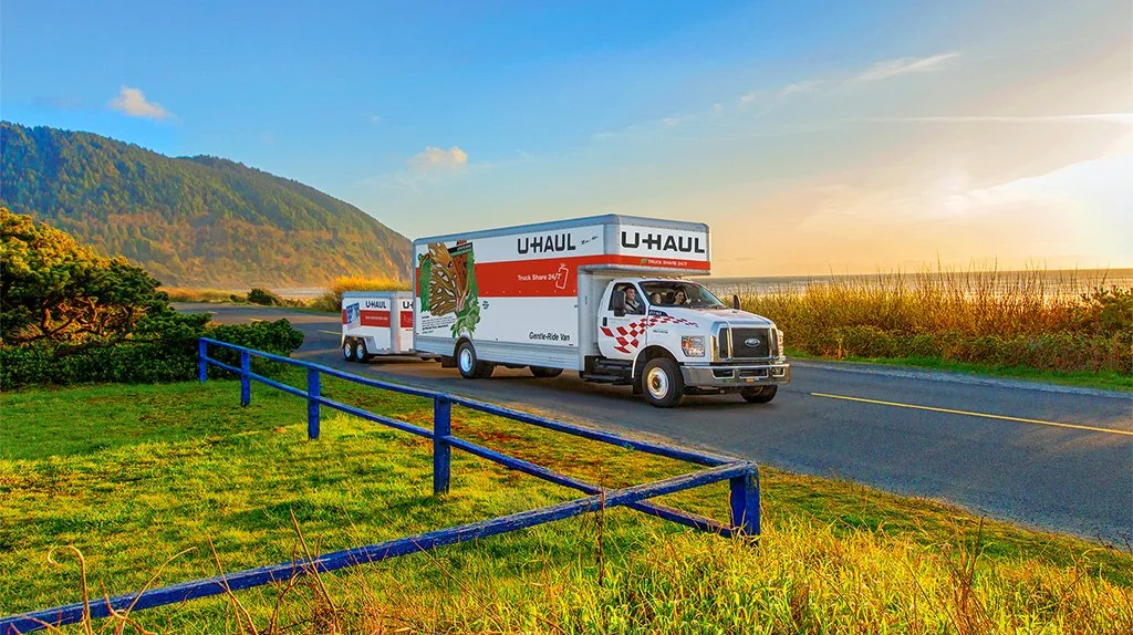 U-Haul trucks traveling along a rural road with mountains and a sunset in the background.