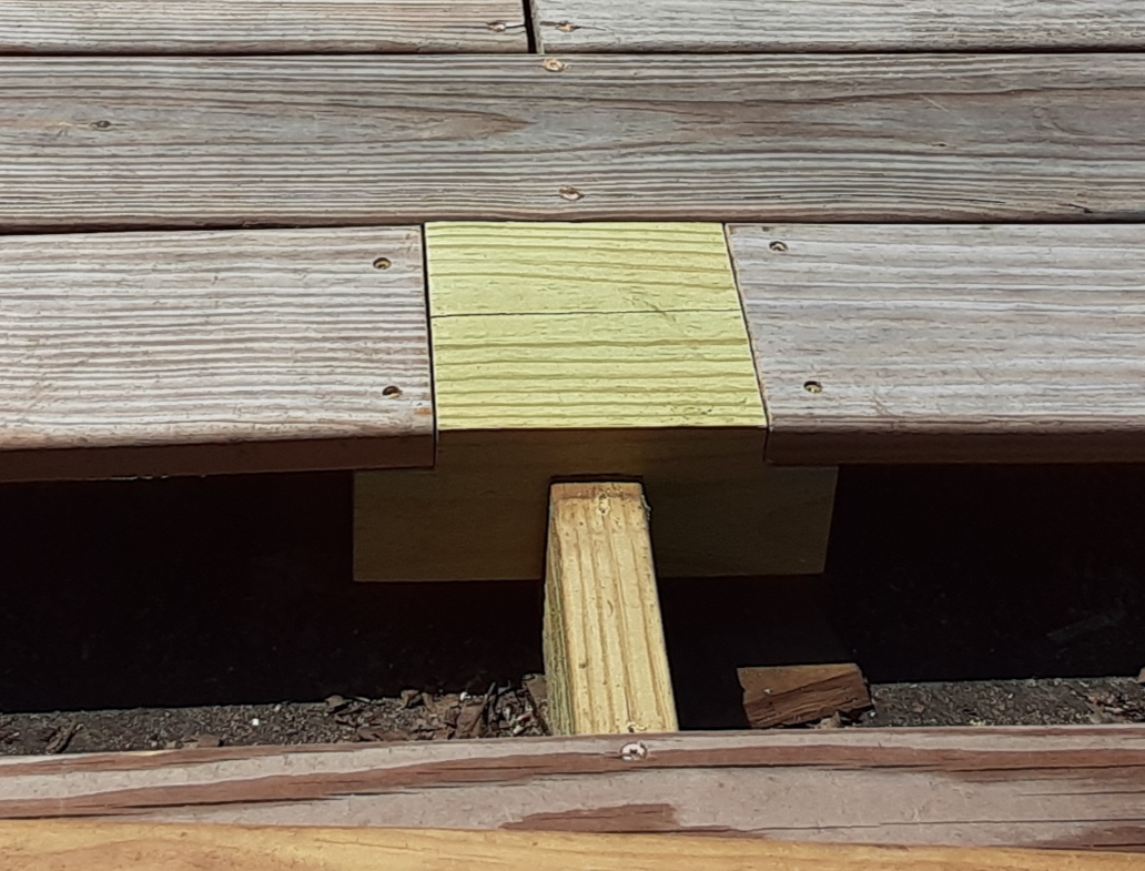 Close-up of wooden deck construction showing a new yellowish wooden block bridging two sections of weathered wooden planks, with a supporting vertical piece underneath.