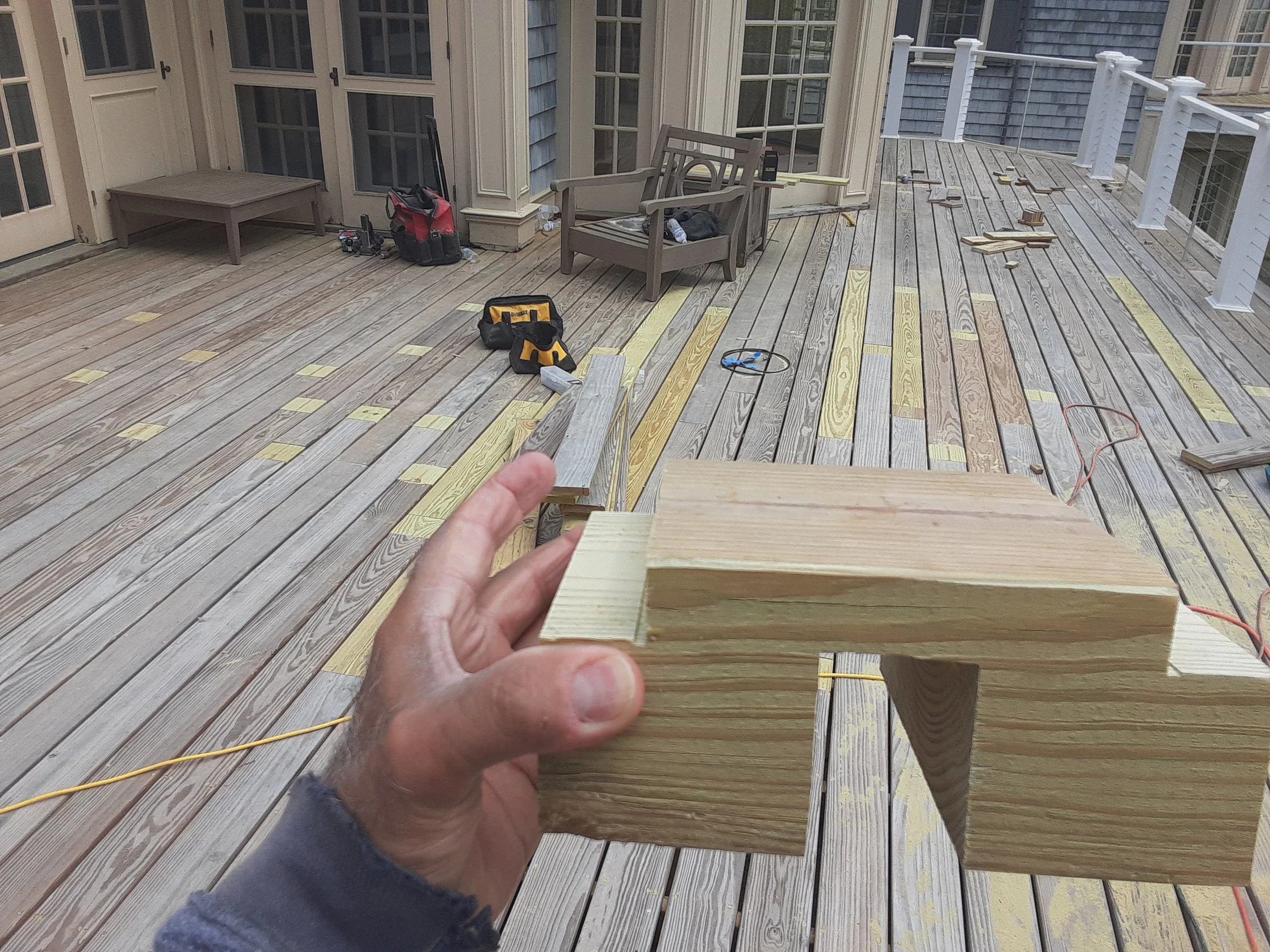 Person holding a small wooden piece on a deck under construction, with tools and wooden planks scattered around.