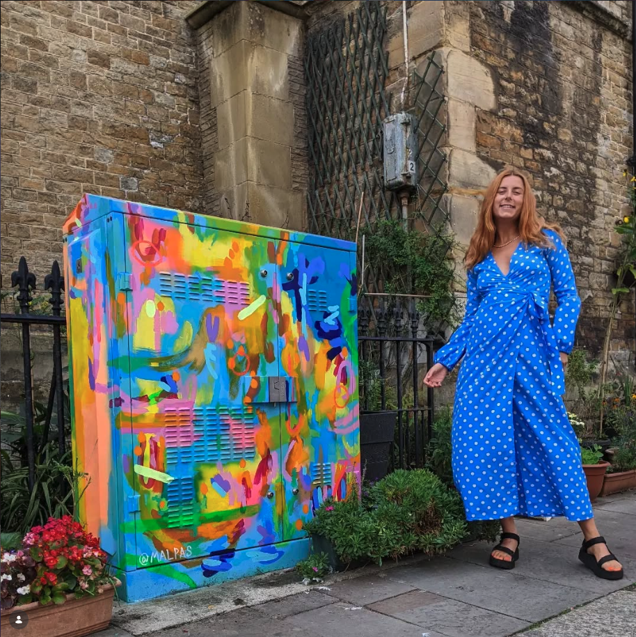 A woman with long, wavy hair wearing a blue polka dot dress and black sandals standing on a sidewalk next to a colorful, abstract mural painted on a utility box, with a brick wall and some green plants in the background.