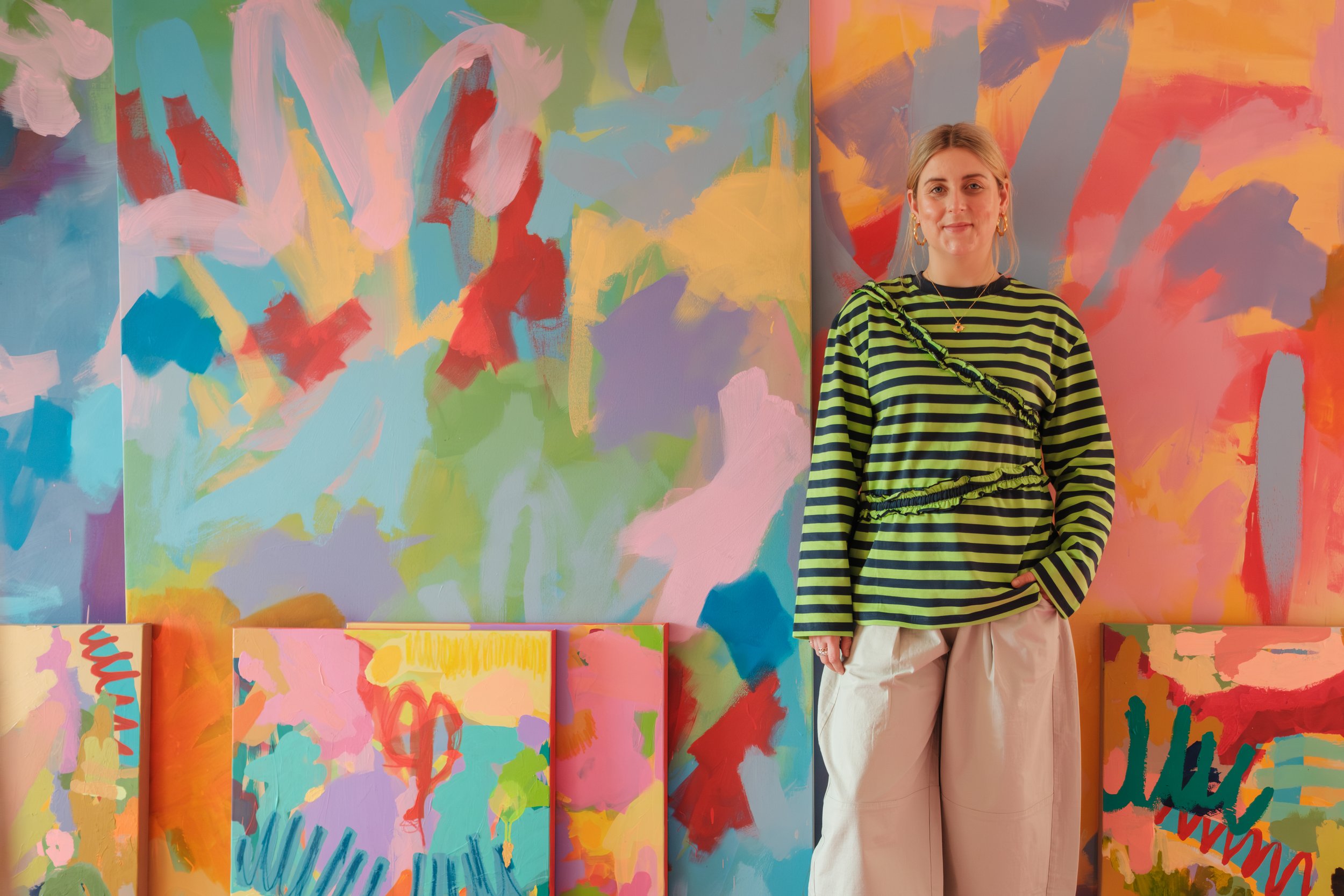 Young woman standing in front of colorful abstract paintings in an art gallery, wearing a green and black striped shirt and beige pants.