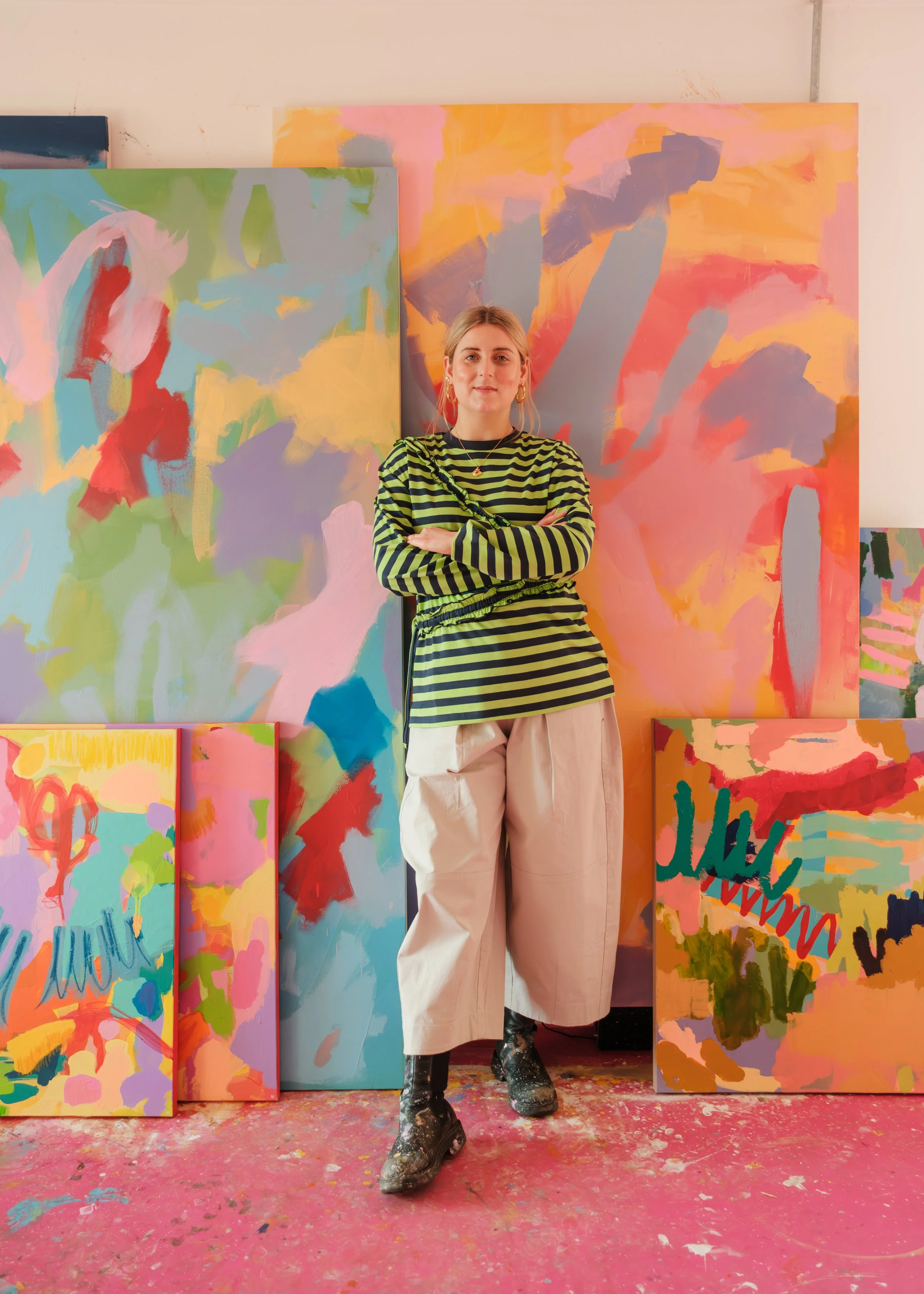 A young woman with blonde hair tied back, wearing a black and green striped long-sleeve shirt, wide beige pants, and black paint-splattered boots, stands with arms crossed in front of colorful abstract paintings in an art studio.