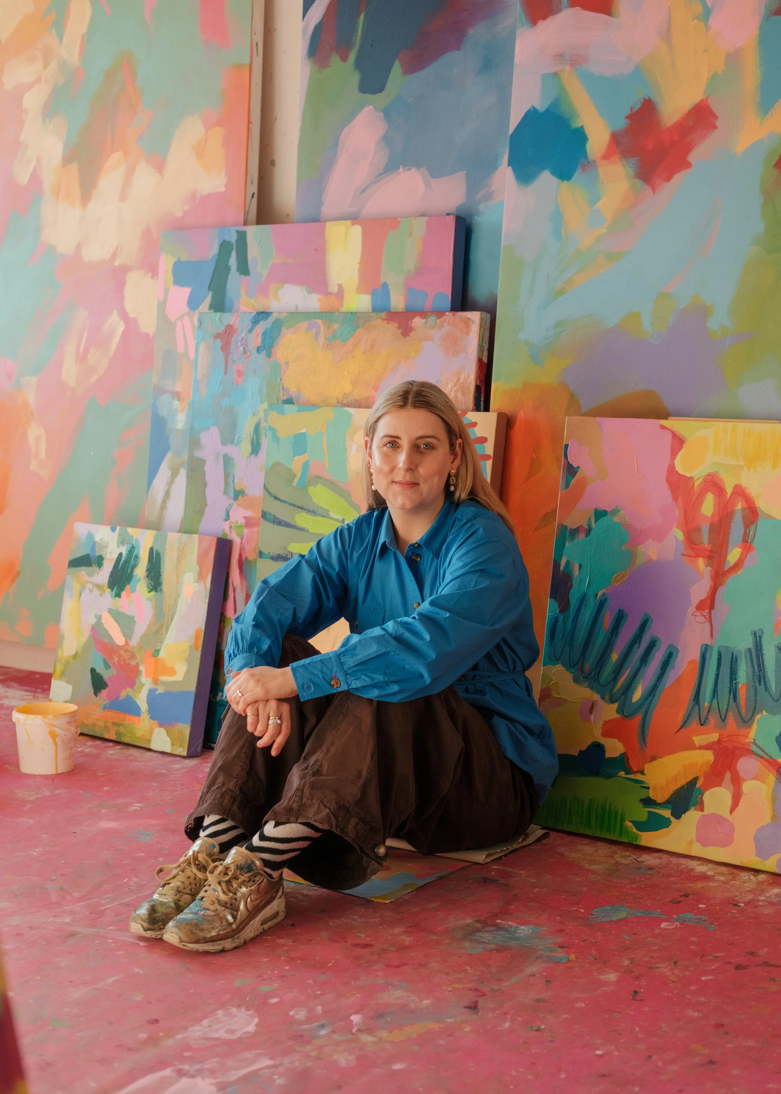 A woman with long blonde hair sitting on the floor in front of colorful abstract paintings inside an art studio.
