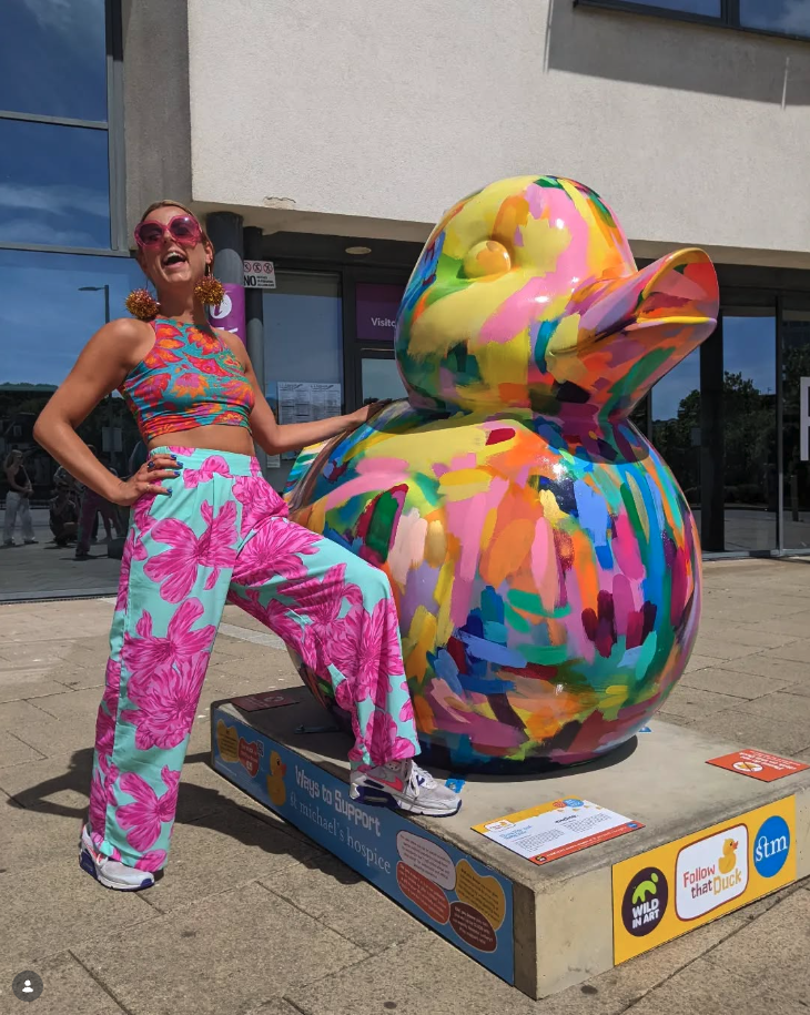 Woman in colorful floral pants and a cropped top standing next to a large, brightly painted duck sculpture.