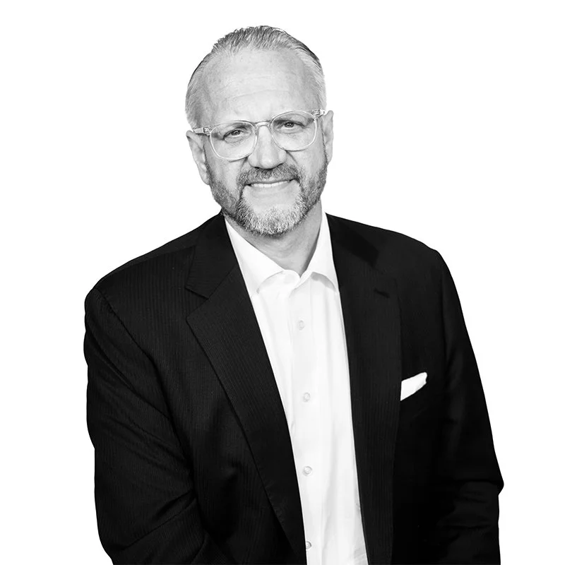 Black and white portrait of a middle-aged man with glasses, a beard, and a friendly smile. He is wearing a dark suit jacket over a white shirt.