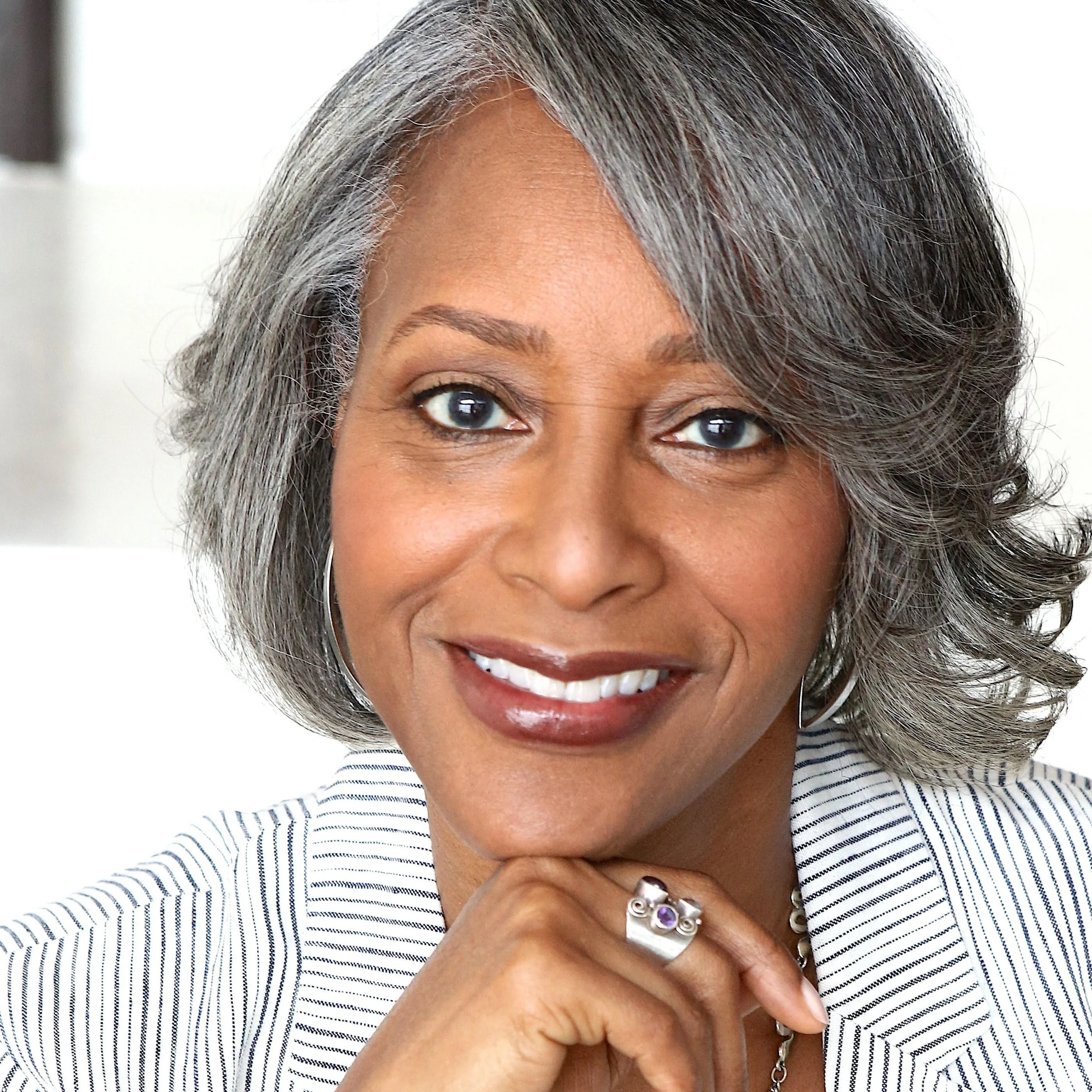 A woman with gray hair, smiling, wearing a striped blazer and jewelry.