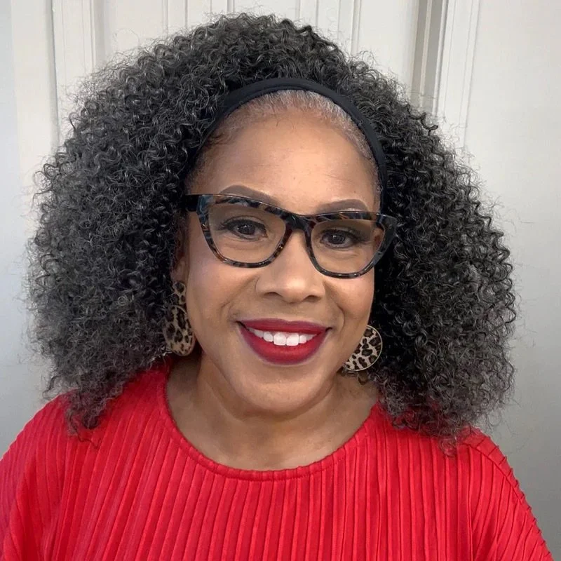 A woman smiling, wearing glasses, red top, leopard print earrings, and a black headband, with curly hair.