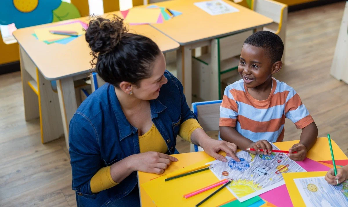 A young boy and a woman sitting at a colorful table in a classroom, coloring a fun Easter-themed picture together.