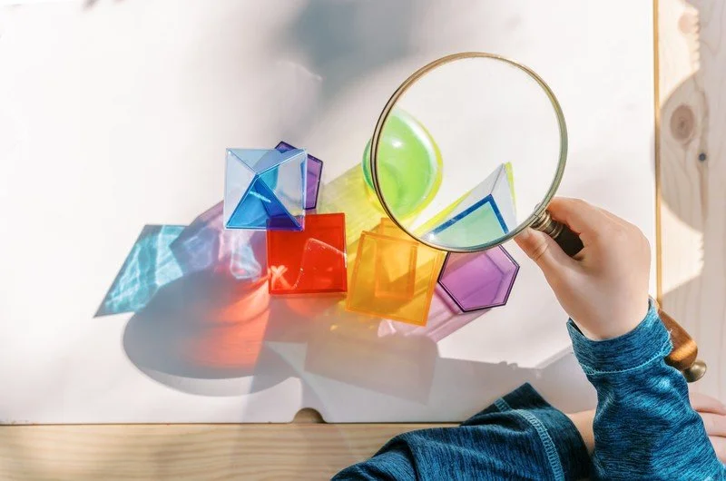 Child holding a magnifying glass over colorful geometric shapes on a white surface.